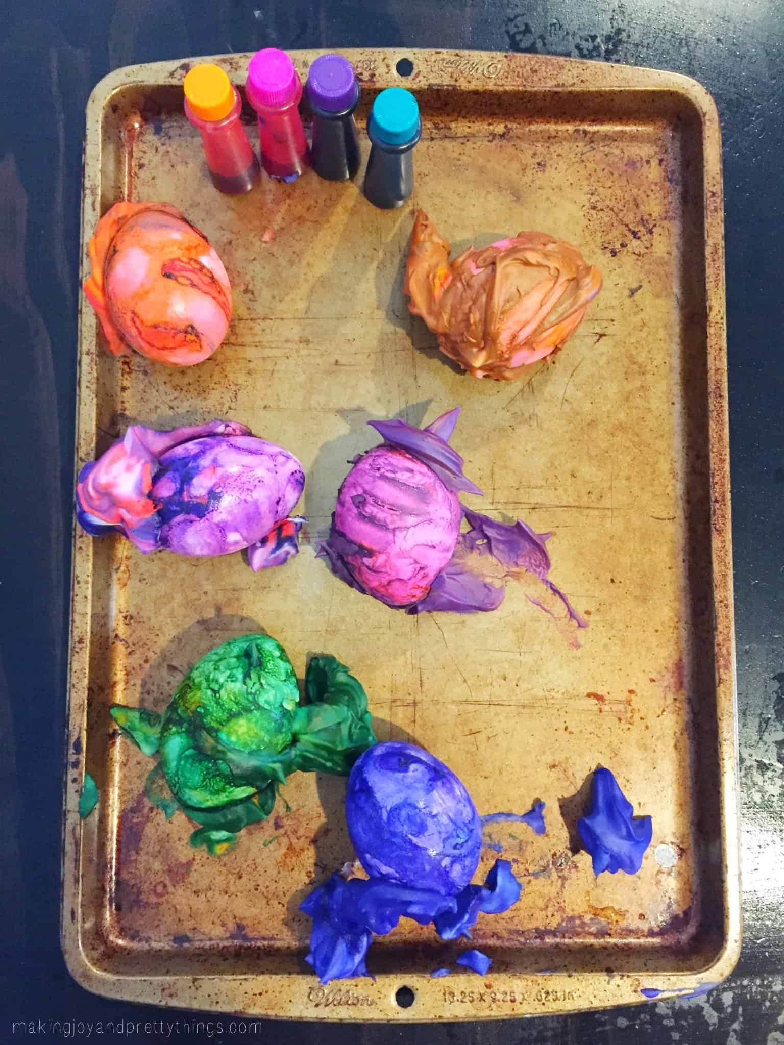 Six eggs covered in blue, green, purple, and pink shaving cream sit on an aluminum baking sheet. The dyed shaving cream covering the eggs has been wiped off, revealing colorful dyed eggs.