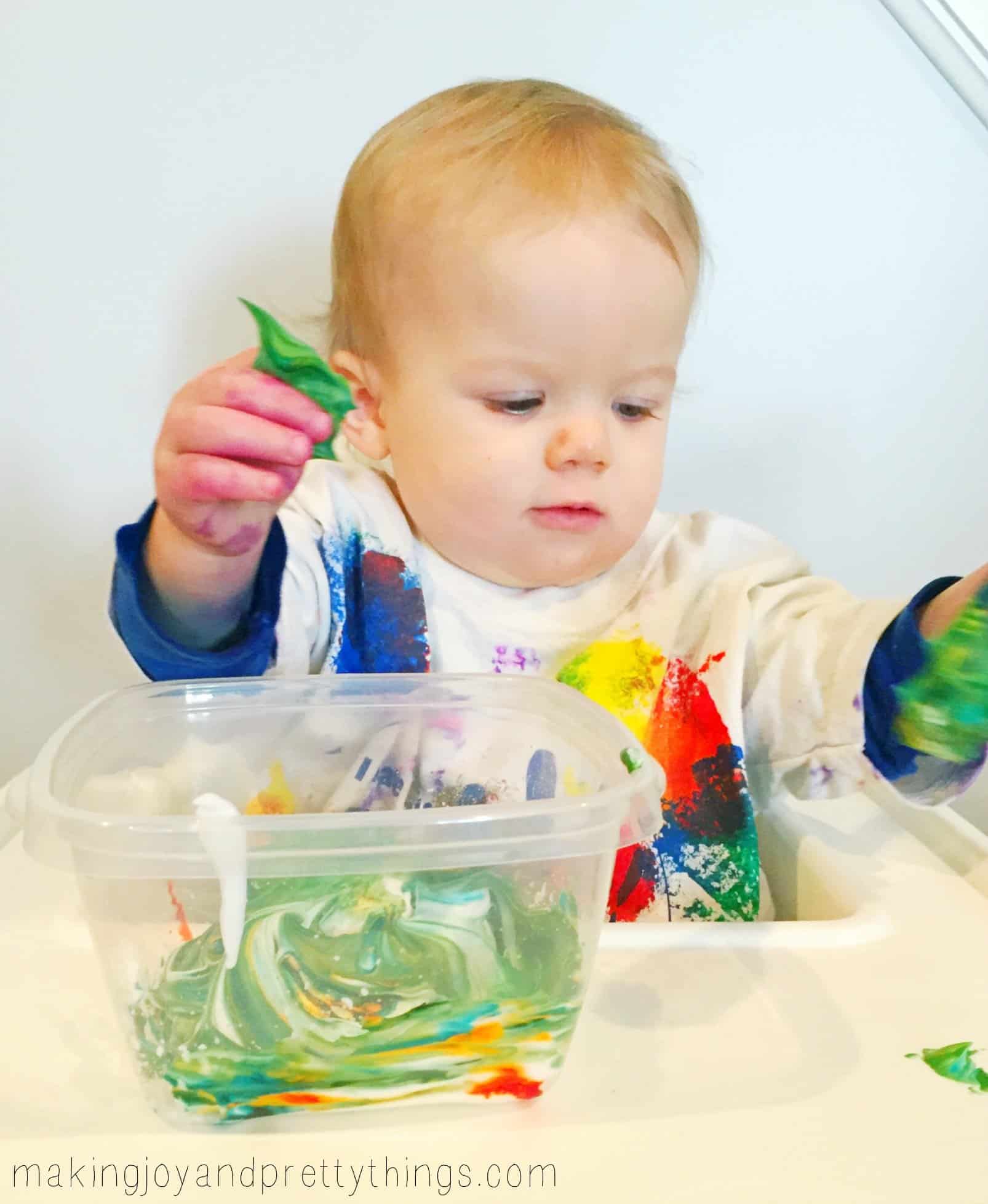 A toddler plays with green-colored shaving cream in a plastic container while sitting in a high chair.