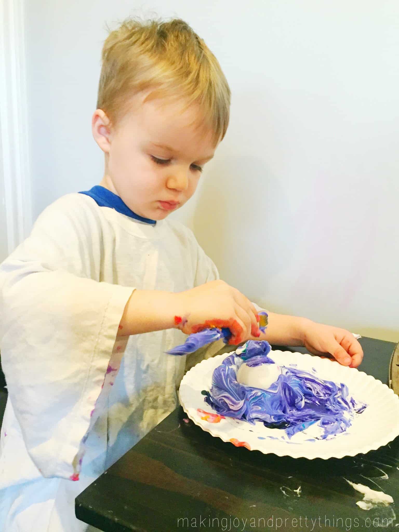  A little boy in an oversized white t-shirt covers an egg with a pile of blue-dyed shaving cream on a paper plate.