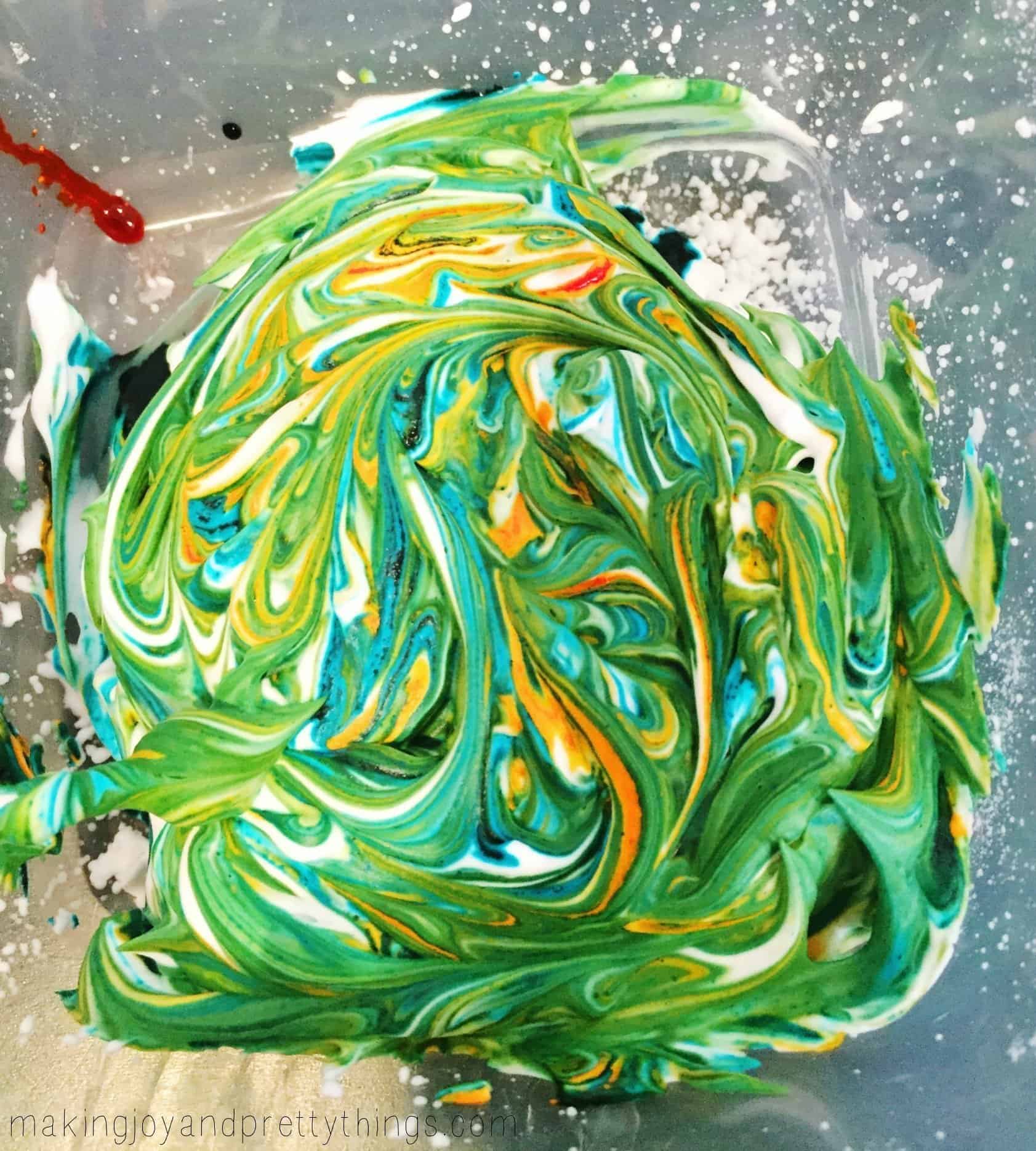 A glass container is filled with a mixture of colored shaving cream. The mixture is a swirl of green, yellow, and blue food coloring.