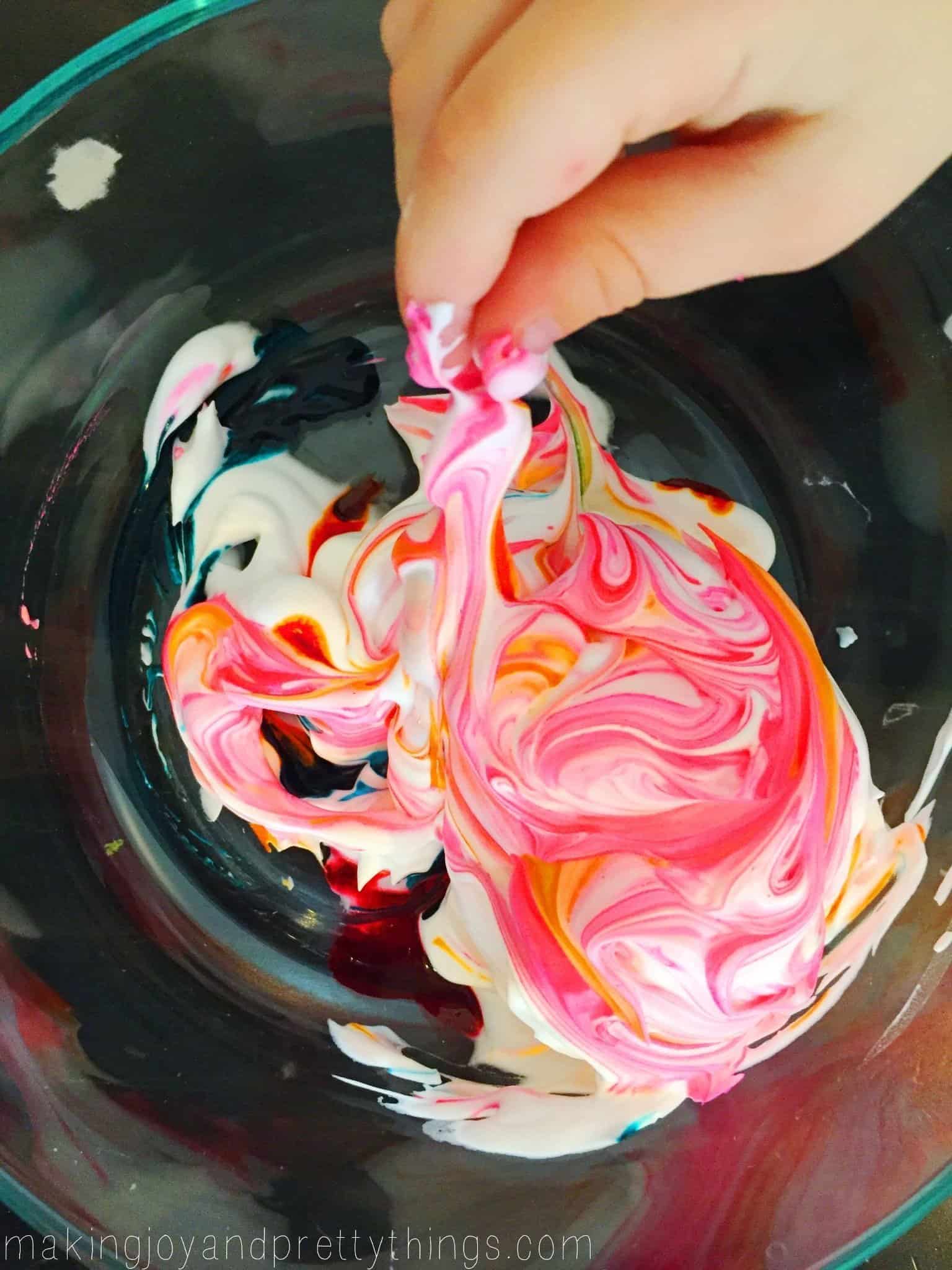 A little boy's hand uses a stick to swirl around pink and orange food coloring into a pile of white shaving cream in a glass bowl.