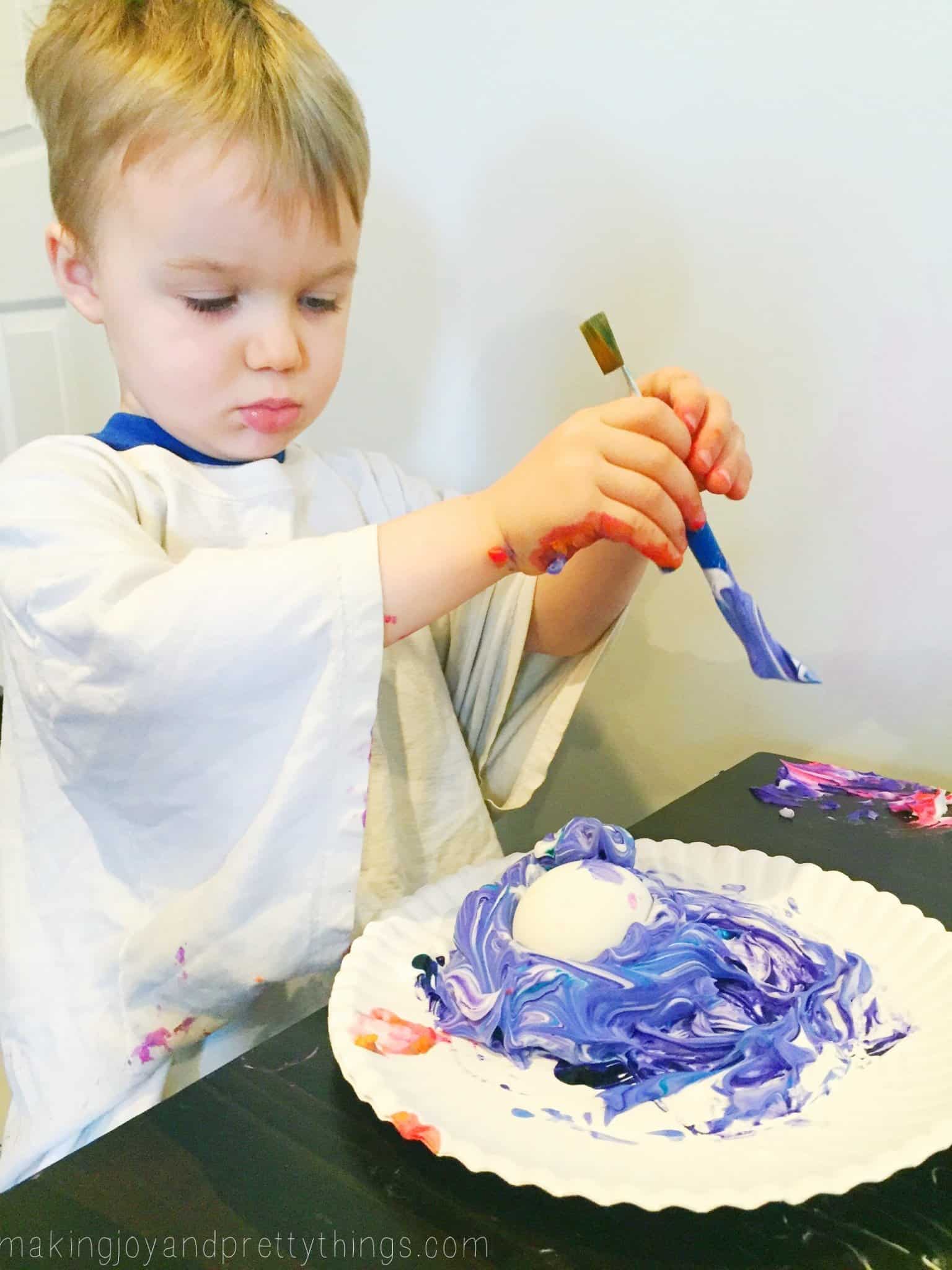 A little boy holds a paint brush and covers a single egg with a pile of blue-dyed shaving cream.
