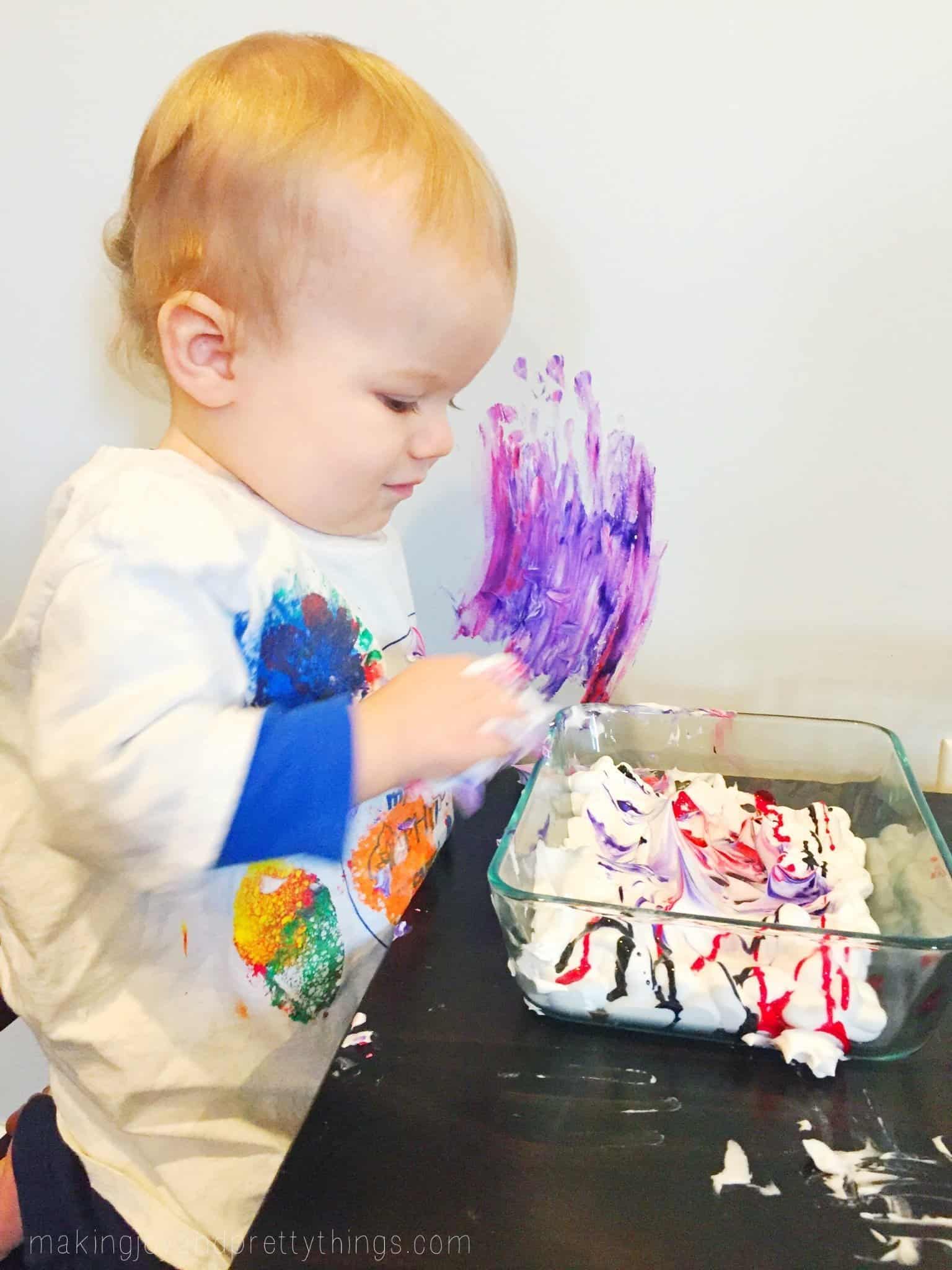 A small toddler sits at a table, a glass dish of white shaving cream mixed with food coloring on the table in front of him. There are streaks of blue and purple hand prints on the white wall next to the table.