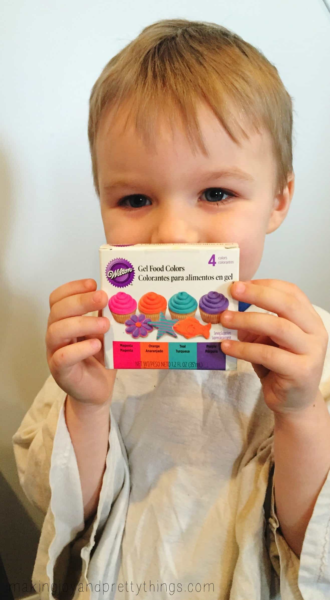 A little boy in a large white t-shirt holds up a box of gel food coloring, which contains for colors - magenta pink, orange, teal, and purple.