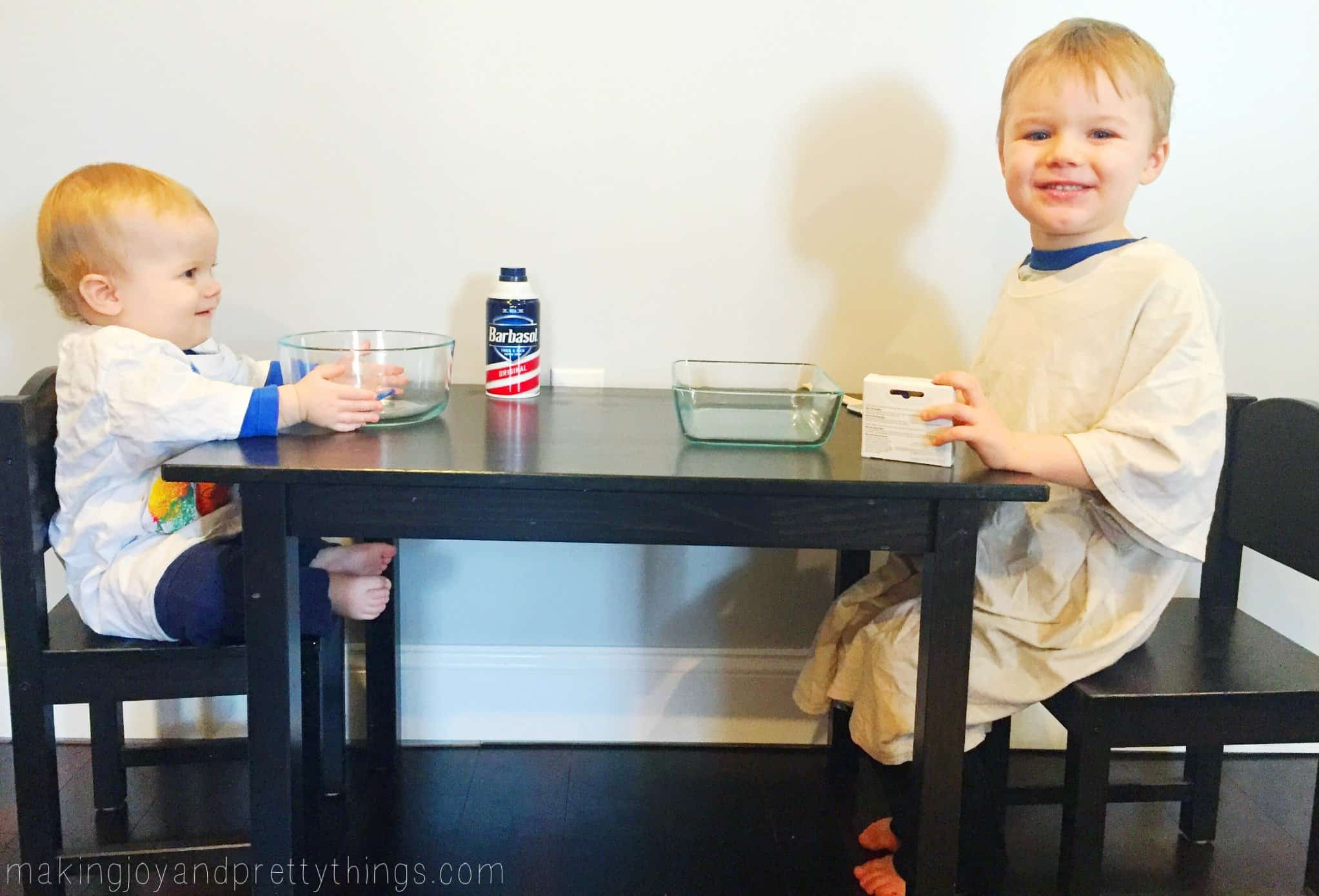 Two young boys sit at a kid-sized table, each wearing an oversized t-shirt and smiling at the camera. On the table are two glass containers and a bottle of shaving cream.