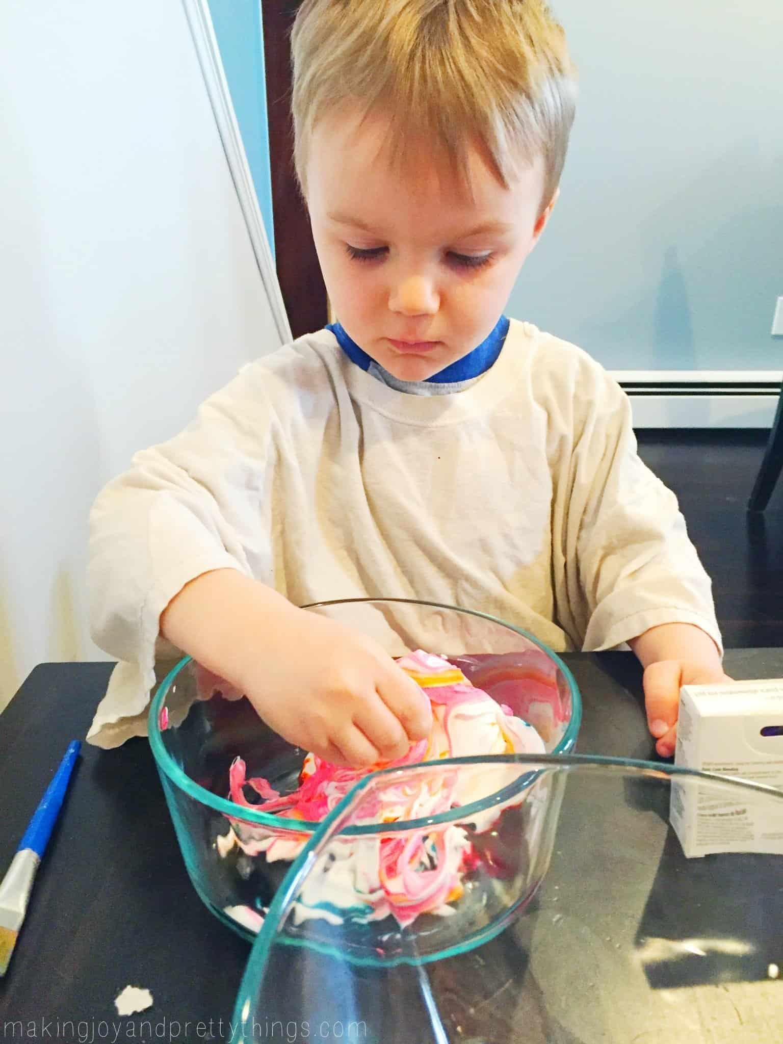 A young boys rolls an egg around in a glass bowl filled with pink and yellow colored shaving cream.