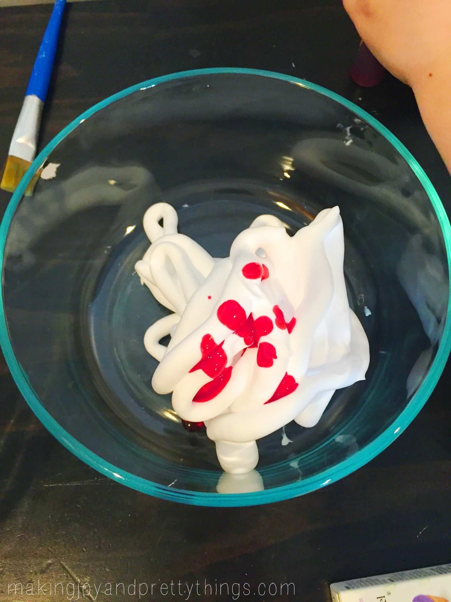 A glass bowl is filled with a pile of white shaving cream, dotted with a few drops of magenta pink gel food coloring.