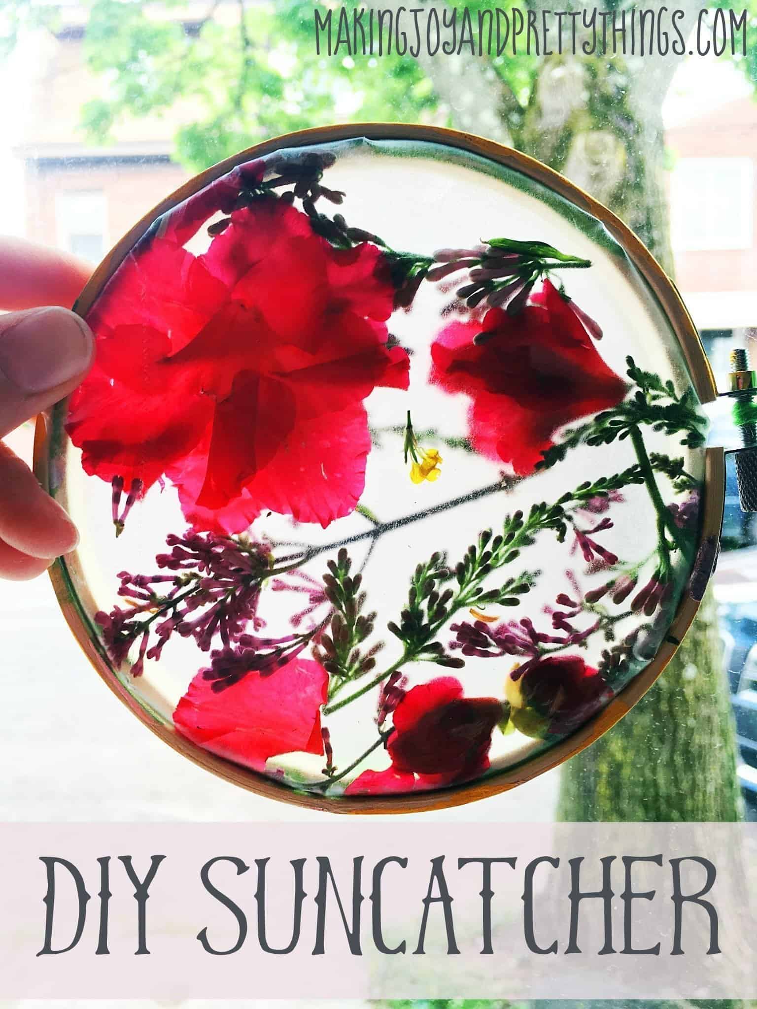 A stunning pressed flower suncatcher is held up in front of a bright window. The transparent suncatcher holds pressed yellow, purple, and pink flower petals in an embroidery hoop.