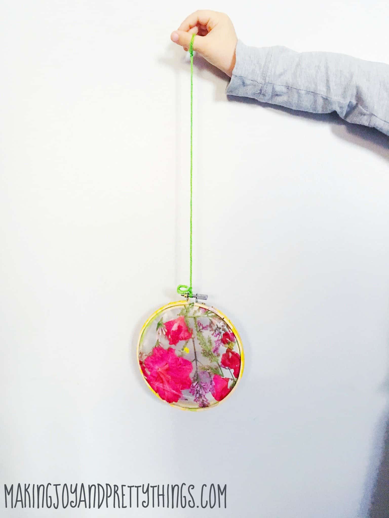 A little boy's arm holds a long piece of green yarn. Hanging on the yarn is a pressed flower suncatcher, made with a yellow-painted embroidery hoop and pink and purple flower petals pressed in contact paper.