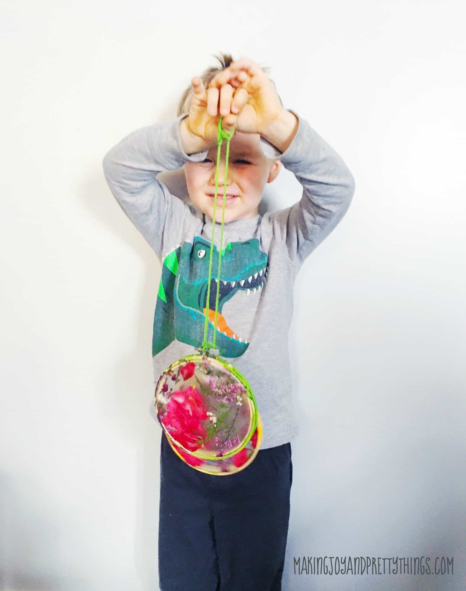 A little boy wearing a gray shirt with a blue dinosaur on it holds up two small pressed flower suncatchers, hanging from two pieces of green yarn. The suncatchers have pink and purple pressed flowers between yellow and green-painted wood embroidery hoops.