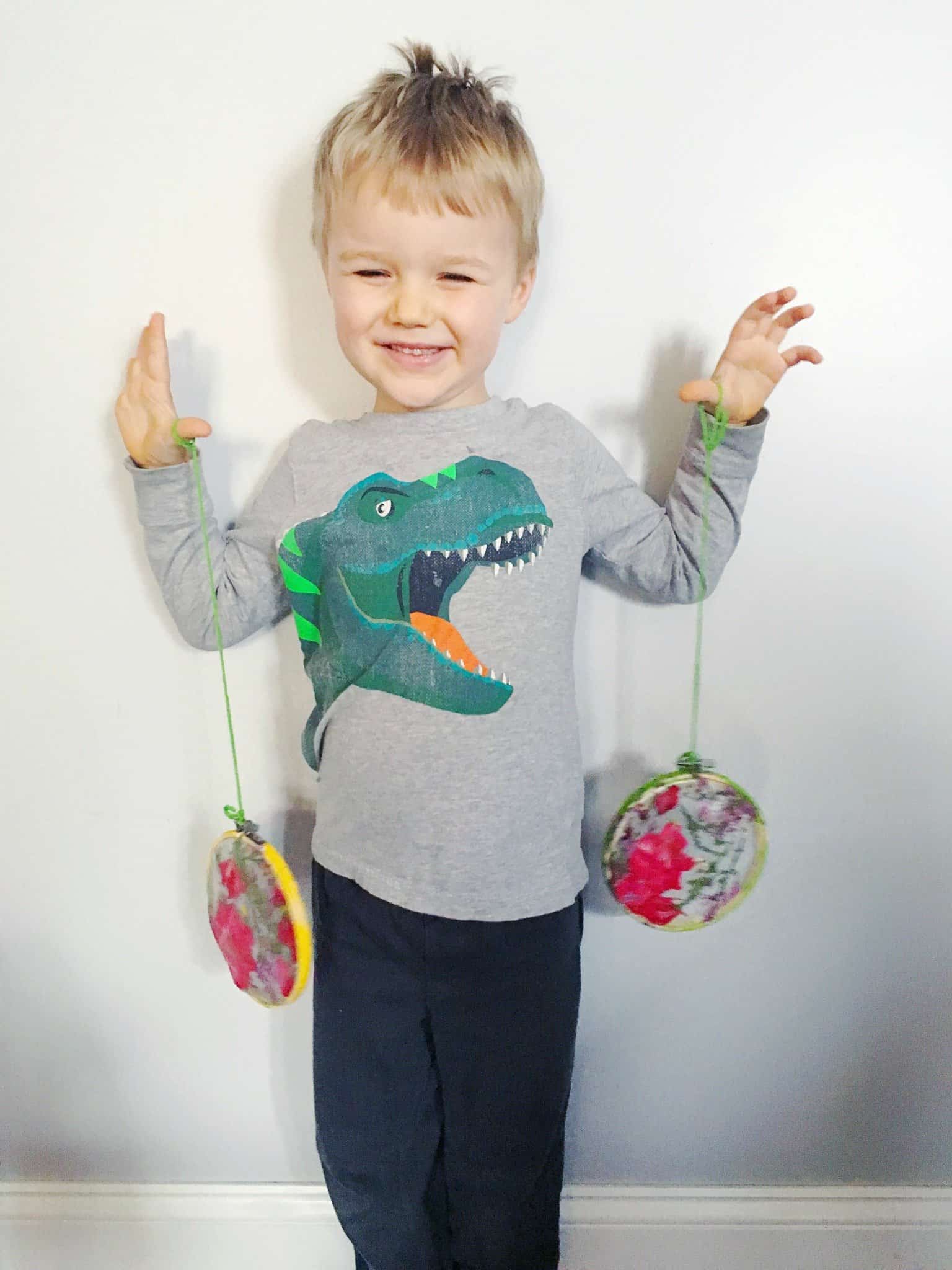 A little boy wearing a gray long-sleeved shirt with a cartoon dinosaur smiles proudly at the camera, holding two pressed flower suncatchers dangling from pieces of green yarn.