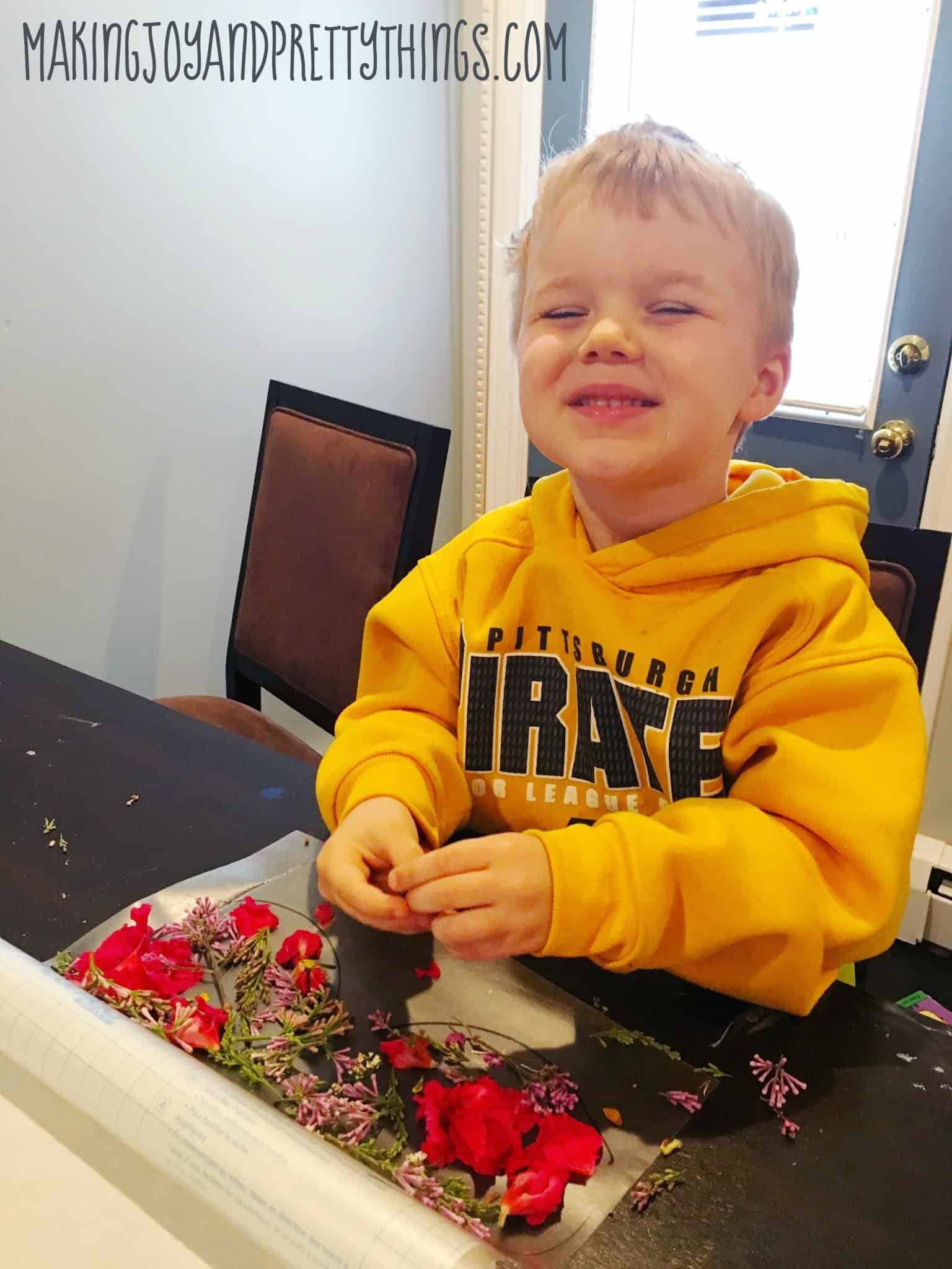 A little boy wearing a yellow sweatshirt smiles at the camera. On the table in front of him is a sheet of contact paper with a variety of pink, purple, and green flower petals arranged in two hand-drawn circles.