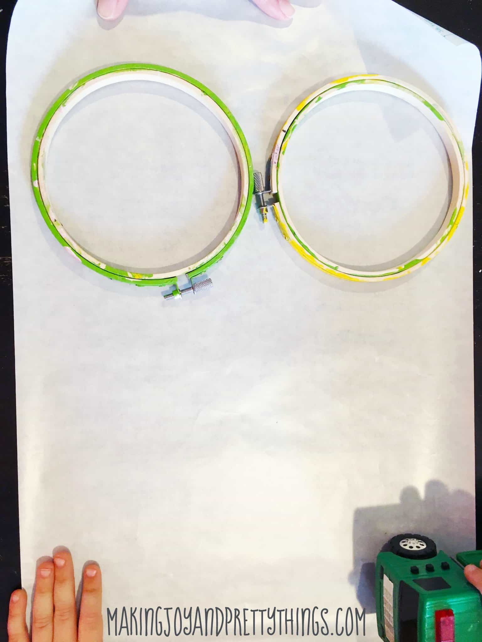 Two small craft hoops, painted yellow and green, sit on a sheet of opaque white contact paper.