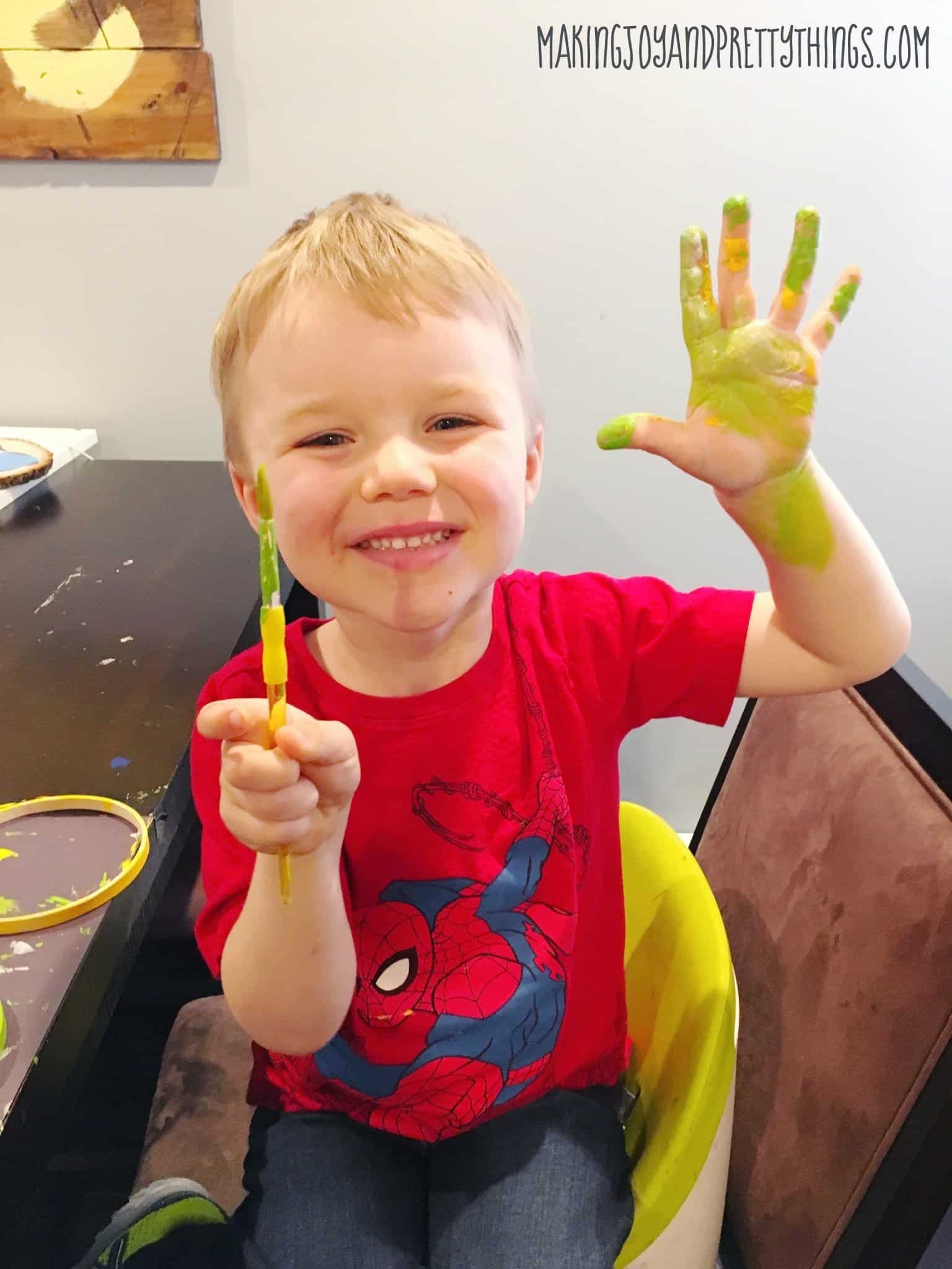 A little boy in a red shirt proudly smiles, holding up his green and yellow paint-covered hand and paint brush while making pressed flower suncatchers.