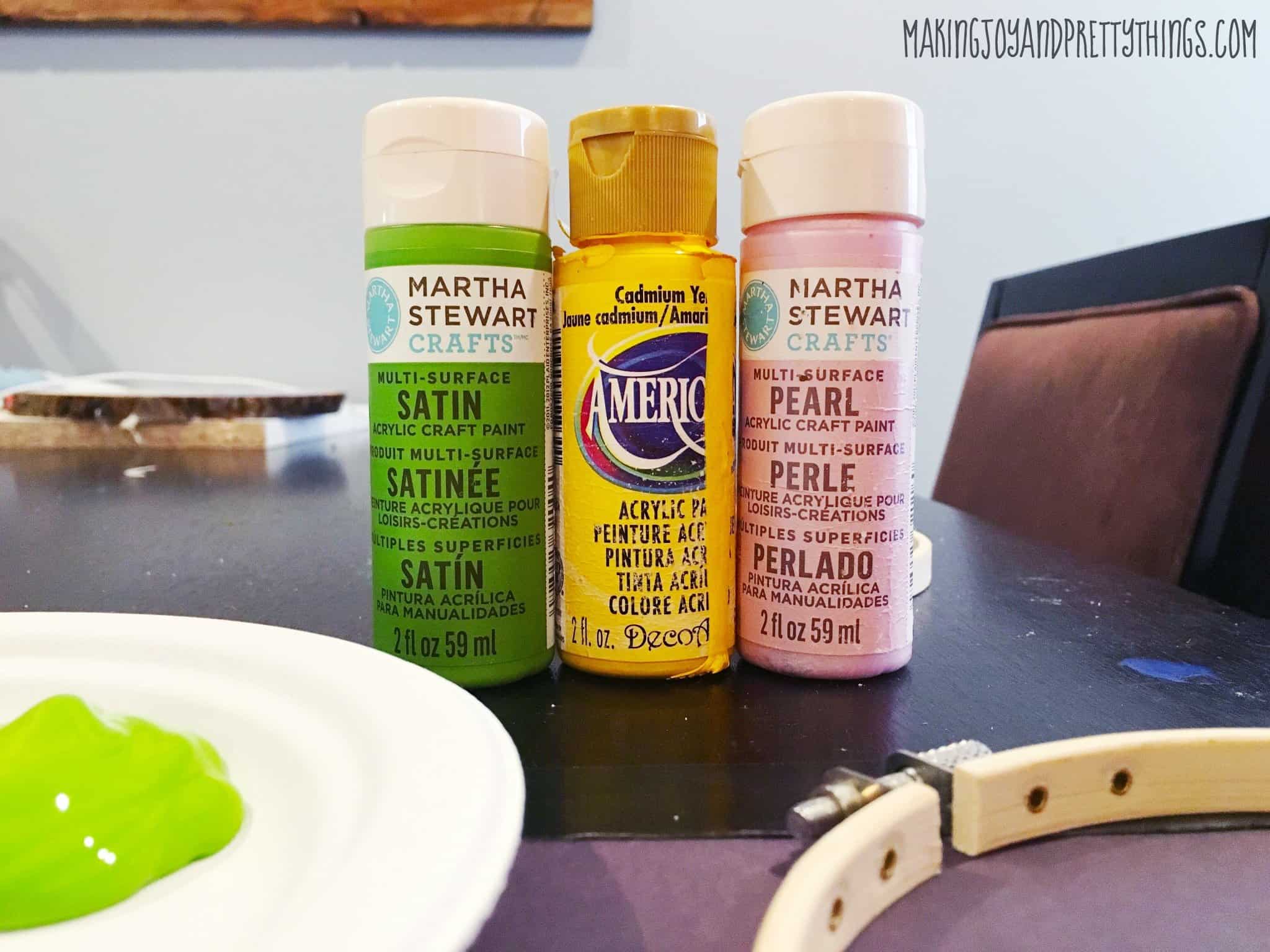 Small bottles of green, yellow, and pink acrylic craft paint lined up on a table. A dollop of green pain sits on a white paper plate, along with an empty craft hoop.