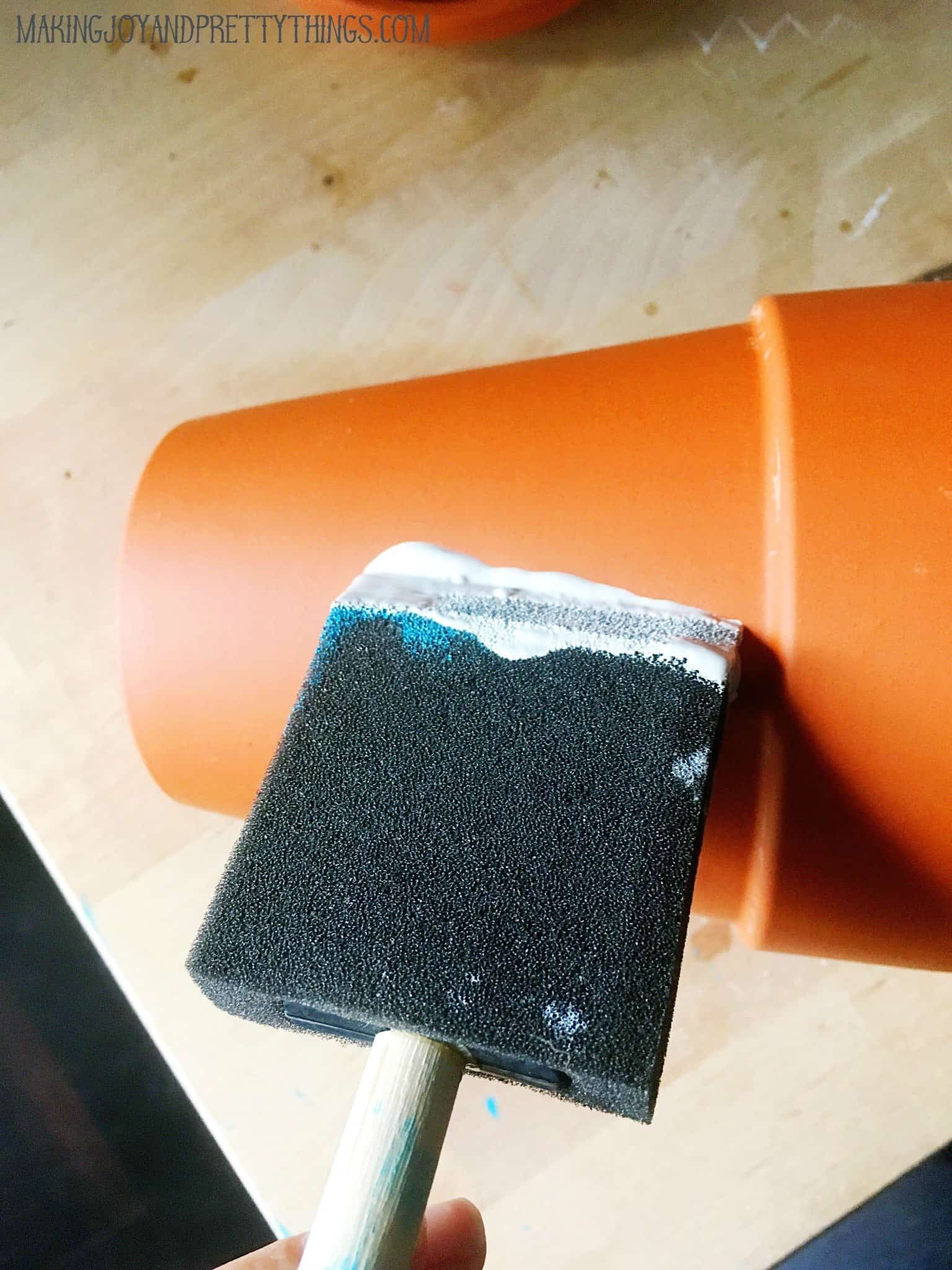 A woman uses a foam crafting brush to paint a terra cotta planter pot with white chalk paint.