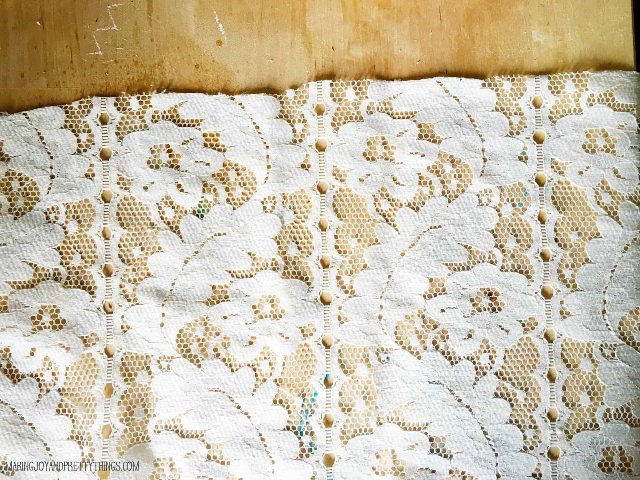 A piece of lace curtain fabric sits on a wood surface, showing a cut line at the top of the fabric.