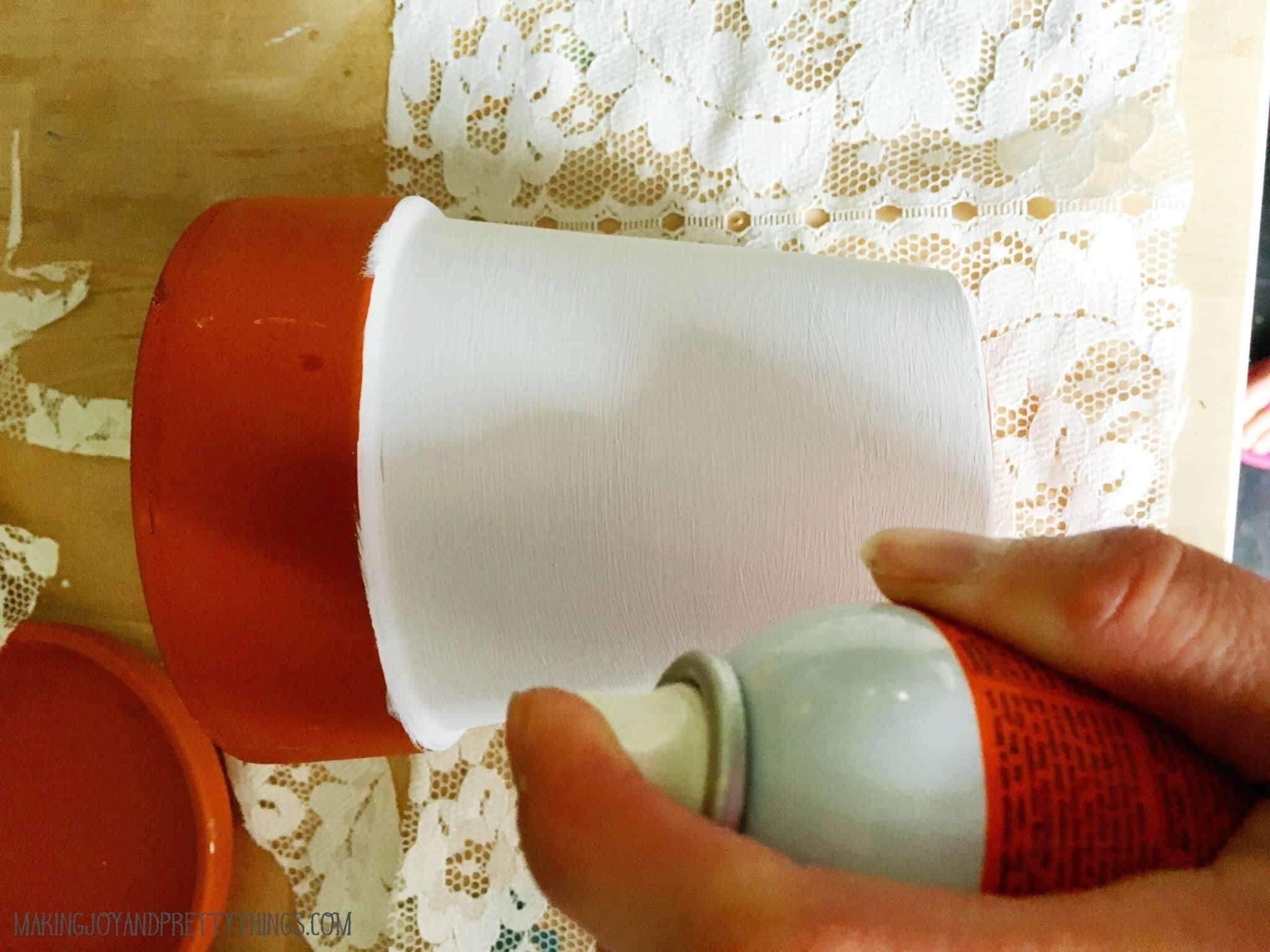 A woman's hand sprays glue adhesive on a partially painted terra cotta planter, which is sitting on top of a piece of lace fabric. 