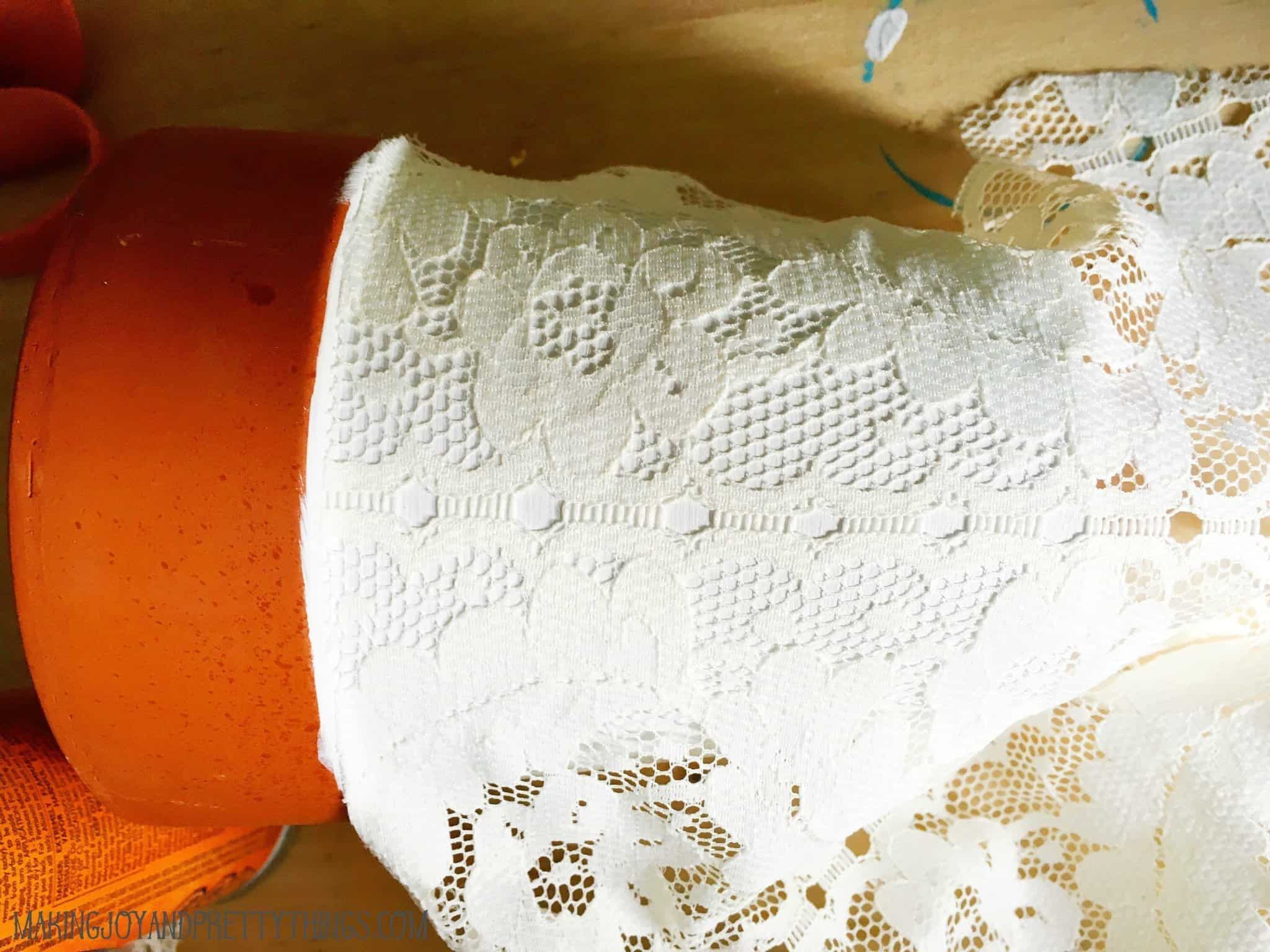 A piece of lace fabric is wrapped around a partially painted terra cotta pot. The lace is wrapped around the white-painted part of the pot, while the natural terra cotta rim remains uncovered.