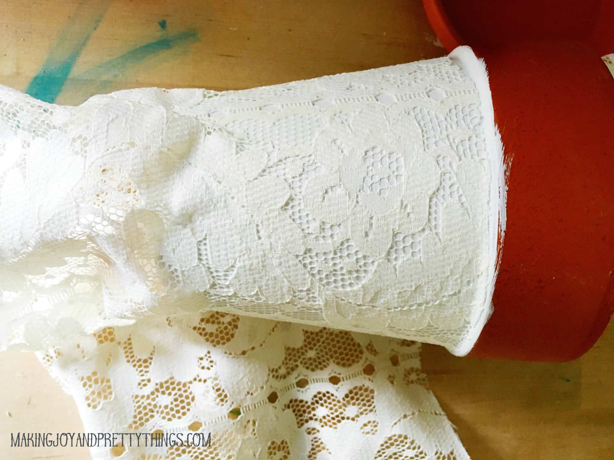 A look at the process of covering a terra cotta pot with lace. Lace fabric is wrapped around a white-painted terra cotta pot. The unpainted rim of the pot remains uncovered.