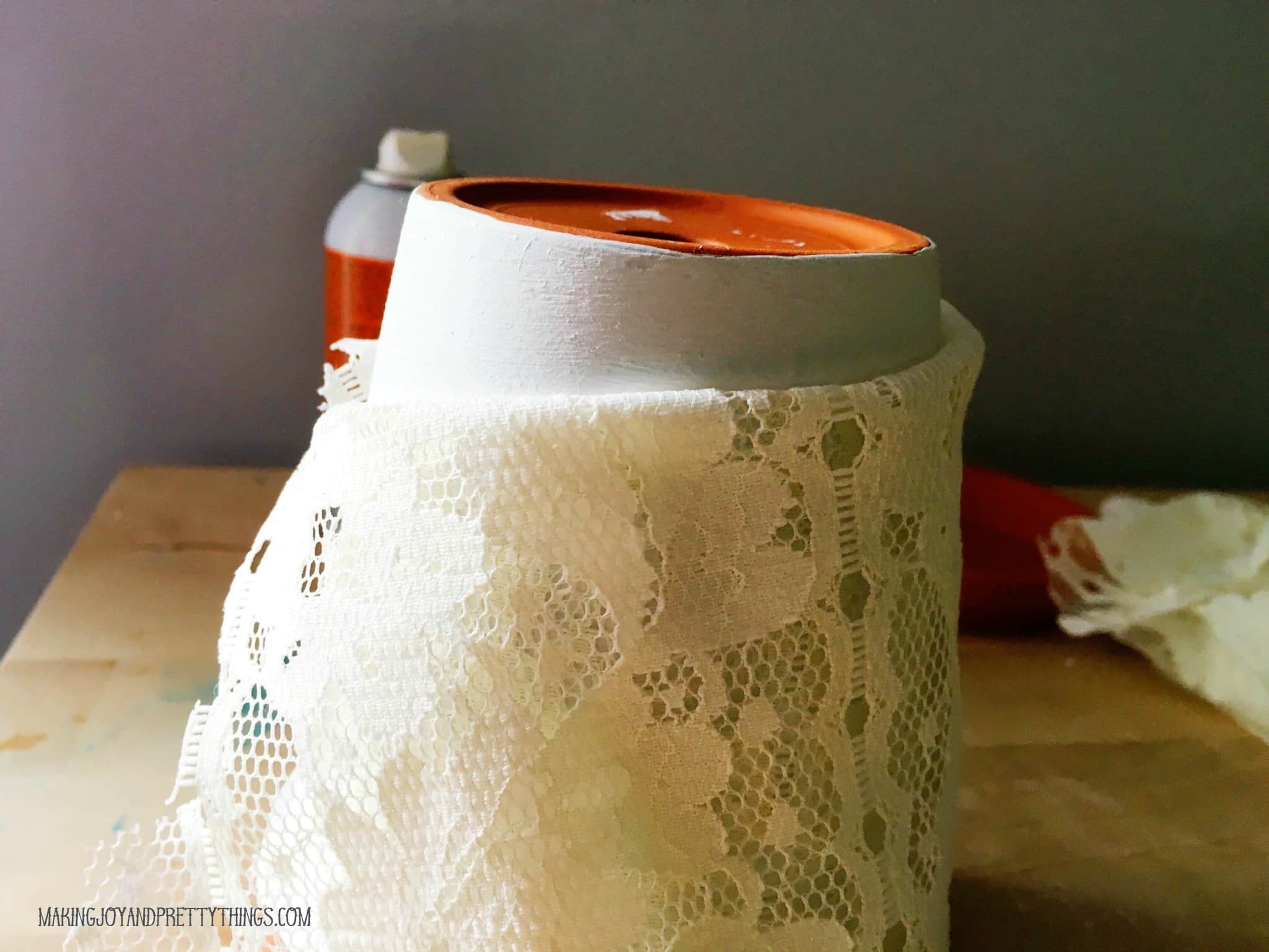 A terra cotta pot is turned upside down with lace fabric wrapped around the body of the pot. The very bottom of the terra cotta pot is unwrapped.