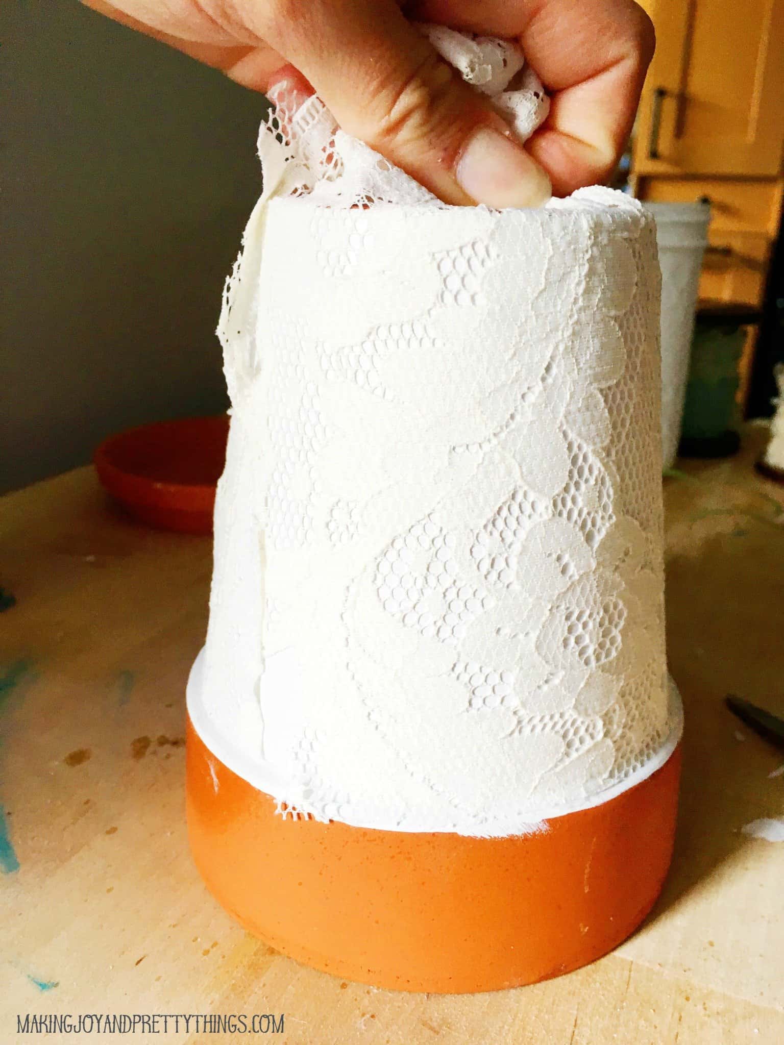 A woman's hand wraps lace fabric around an upside-down terra cotta pot. The pot is painted white, and the lace fabric is bunched at the bottom of the pot.