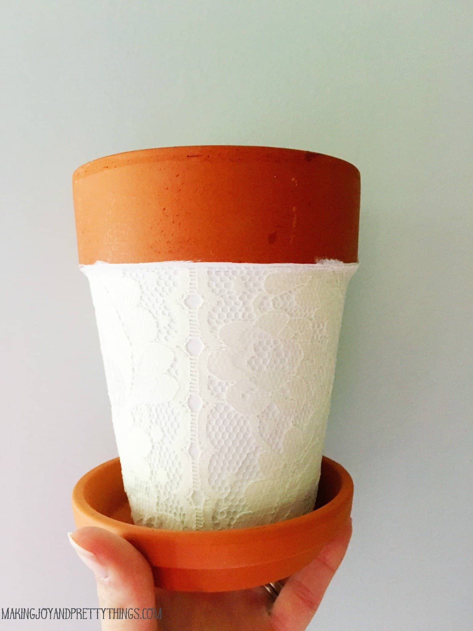 A completed terra cotta pot wrapped in lace fabric. The body of the pot is painted white and wrapped with lace, while the top rim and the saucer remain unpainted.