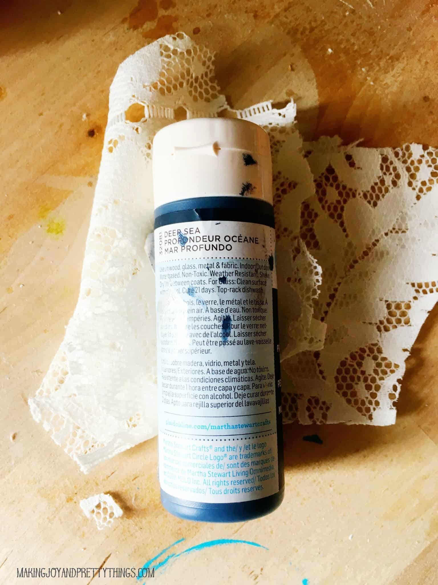 A bottle of dark blue craft paint sits on a swath of lace fabric.