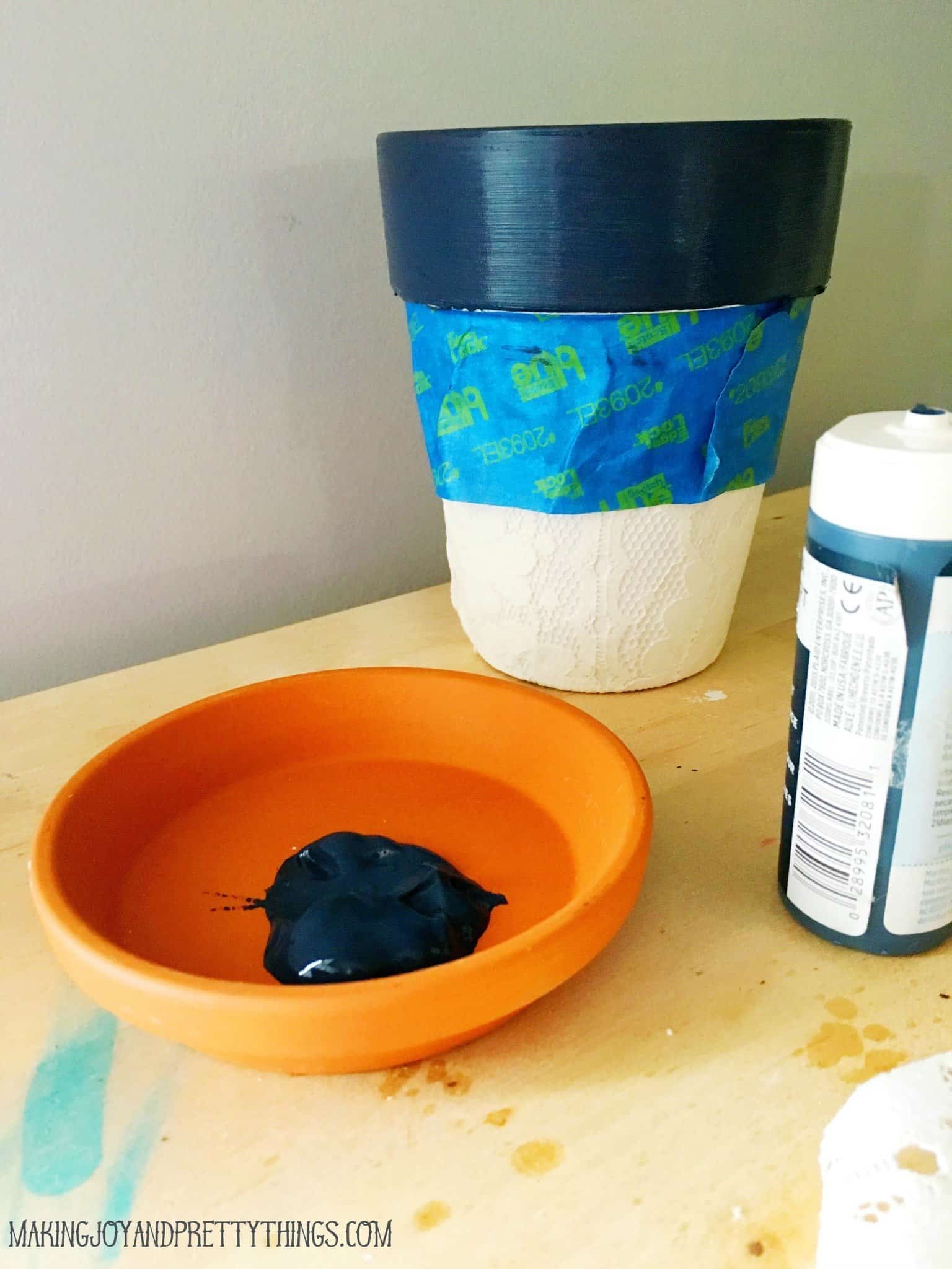 The process of painting a lace covered terra cotta pot. In the background, the pot's rim has been painted a navy blue color, and painter's tape covers the lace body. In the foreground, the terra cotta saucer is being painted with the same dark blue craft paint.