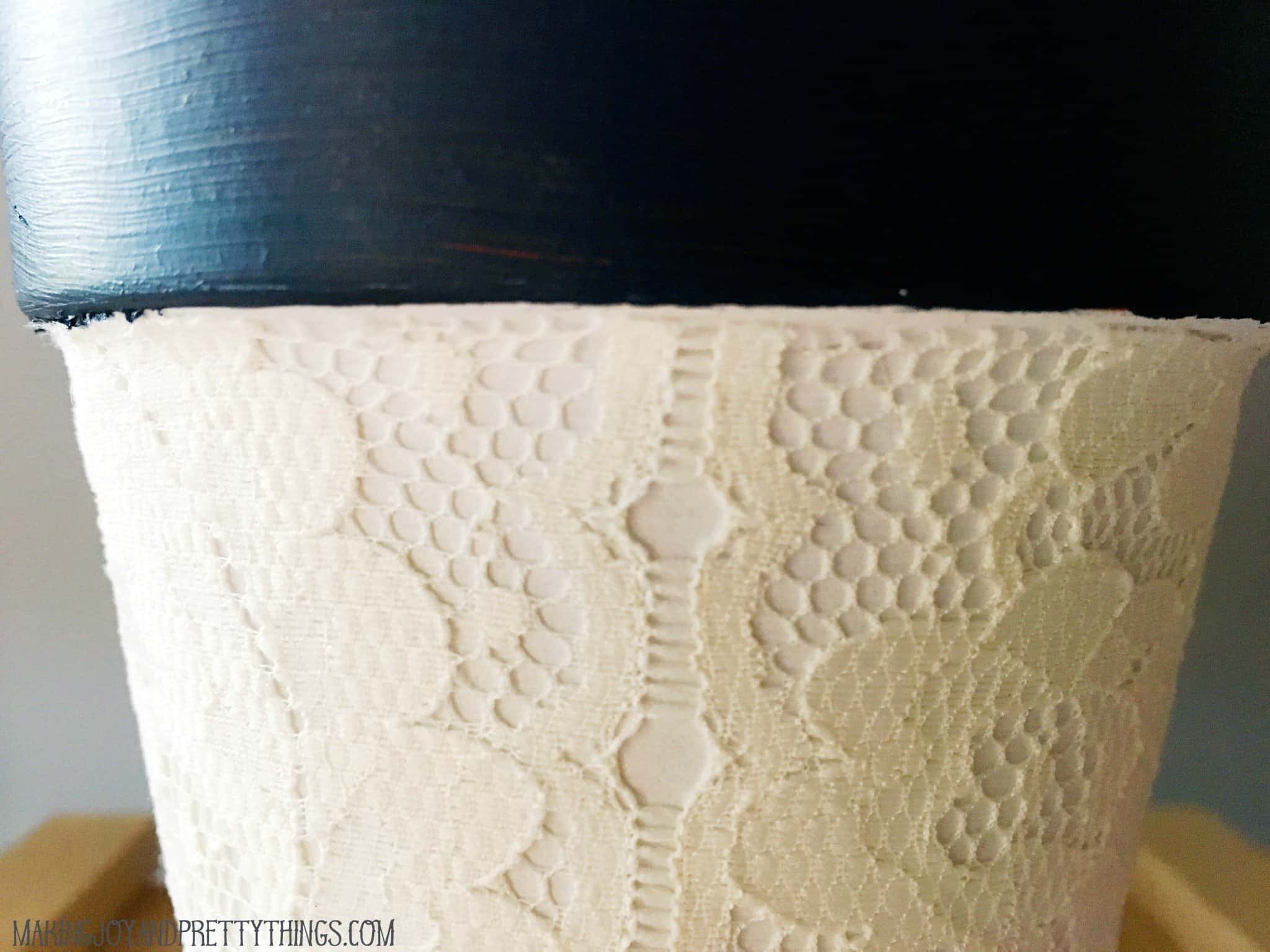 A close up look at the details of the lace fabric covering a white-painted terra cotta pot. The pot's rim is painted a rich, deep blue.