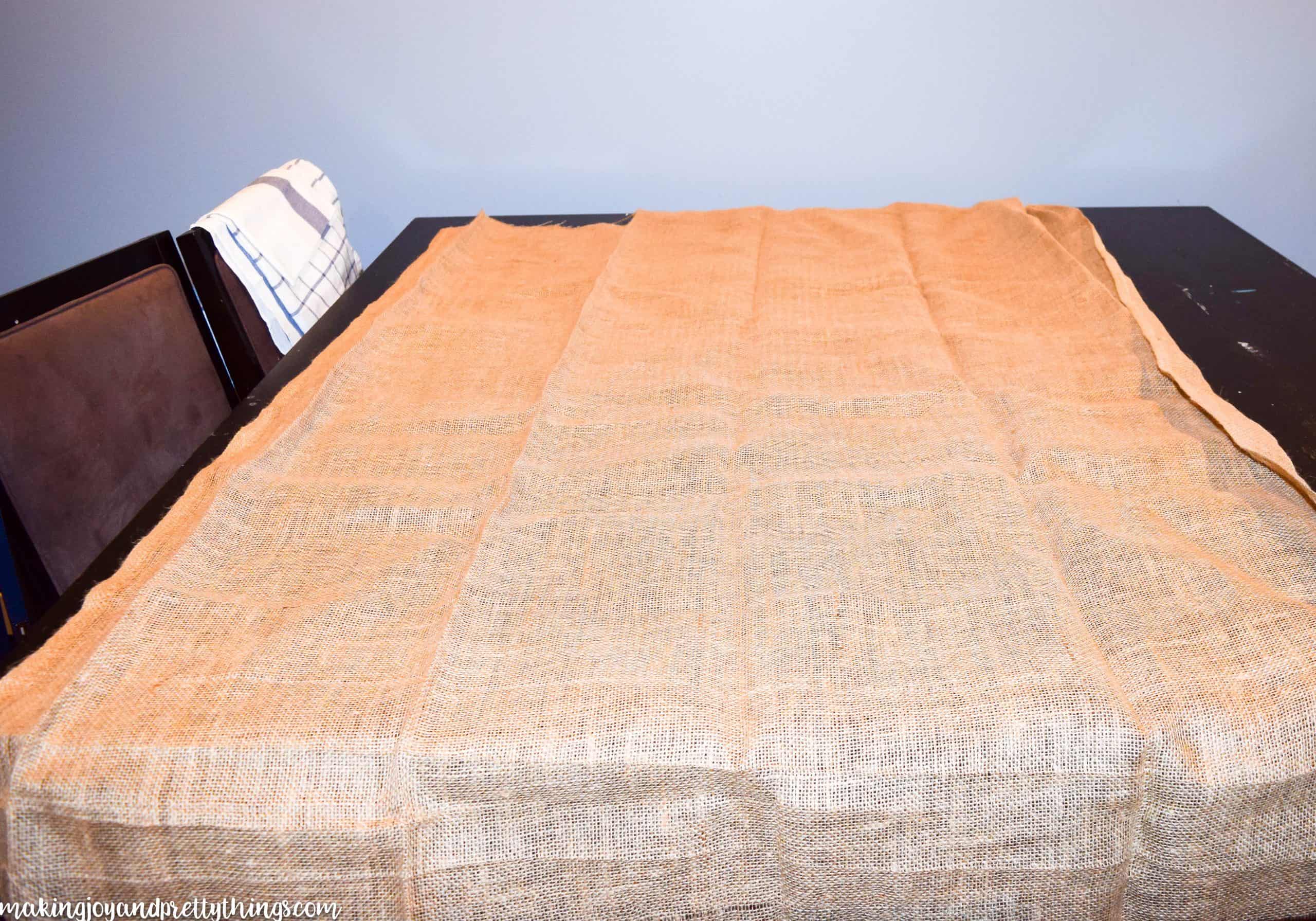 A large sheet of burlap fabric is laid out on a black table. The burlap has creases where it was previously folded.