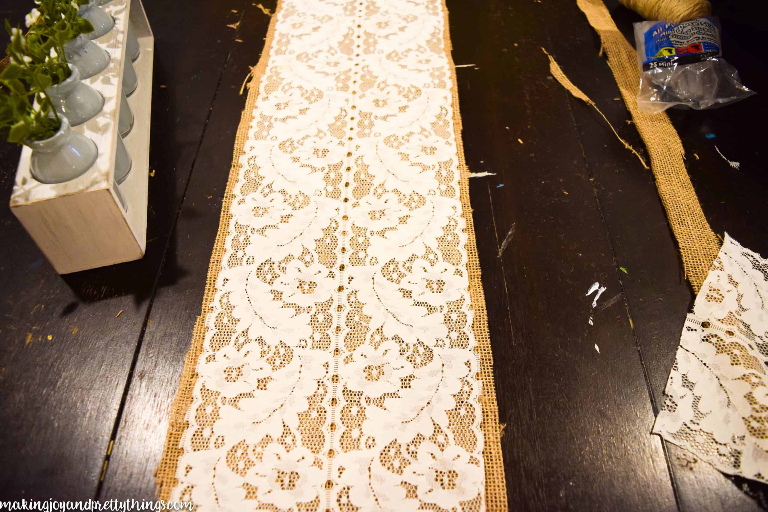 A strip of white lace fabric sits on top of a strip of burlap fabric to create a farmhouse style table runner across a dark wood table.  Scraps of cut fabric surround the runner.