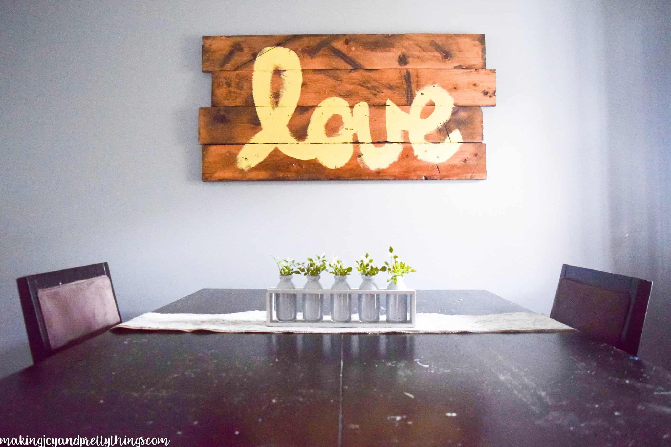 A zoomed-out look at the simple summer centerpiece on a dining room table. The white wood box -filled with five glass jars, each holding sprigs of faux greenery - sits on a lace table runner. In the background, a wood sign hangs on the wall with the word "love" painted across the front.