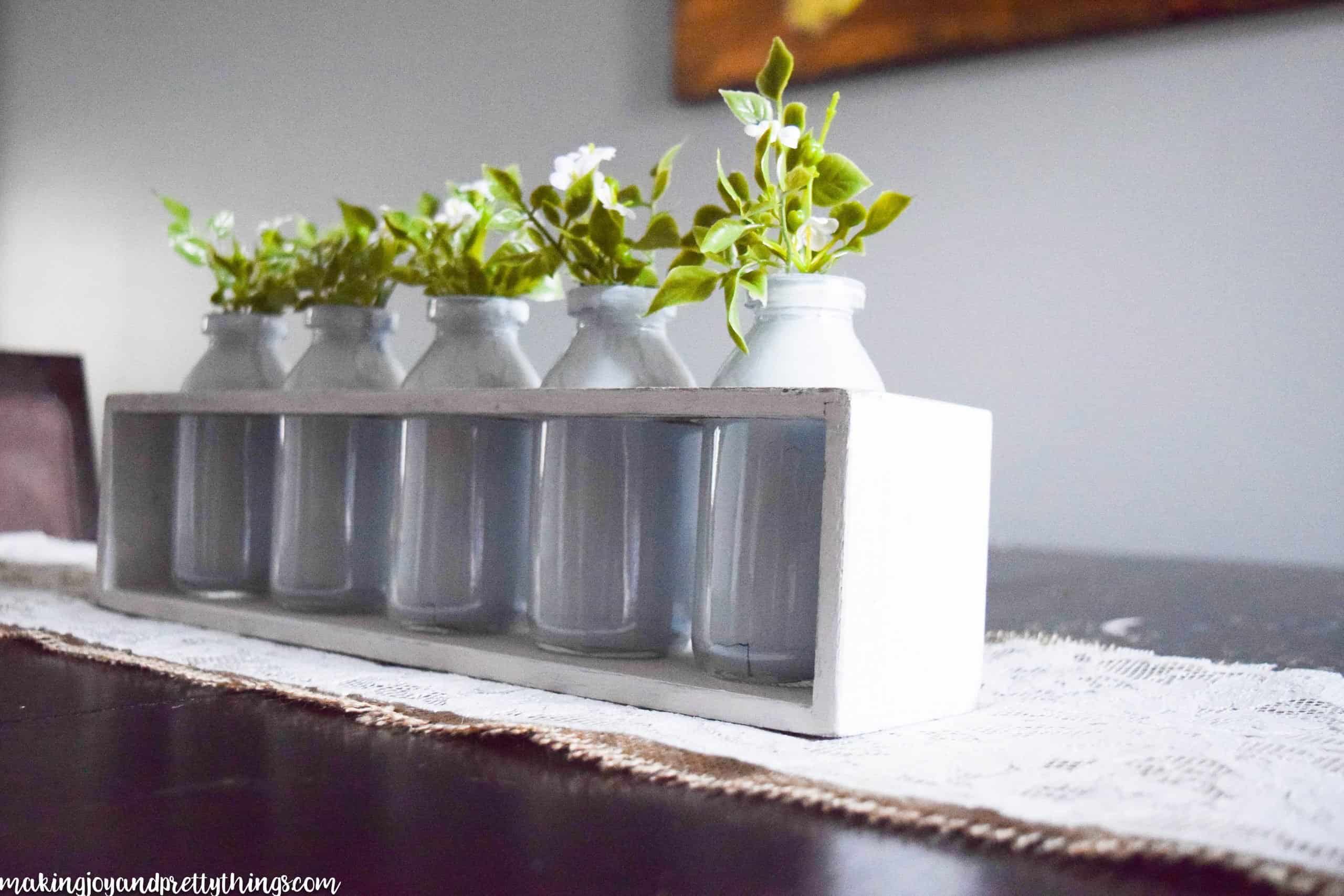 Glass milk jars are lined up in a white-painted wood box. The jars have faux greenery sprigs sticking out from the top.