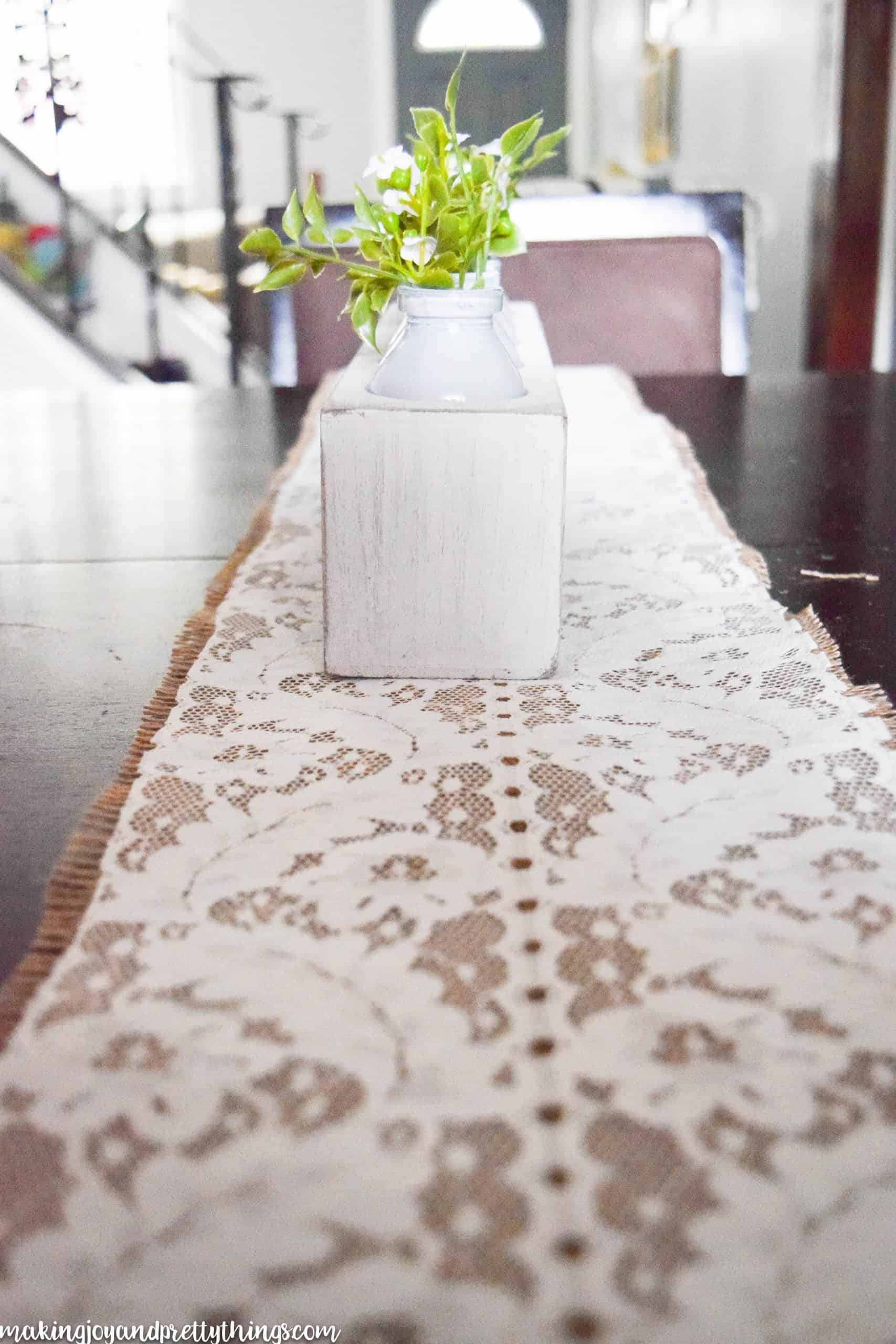 A side-angle view of the summer centerpiece and lace and burlap table runner. Lace fabric sits on top of frayed burlap. A white box with glass jars filled with faux greenery and flowers sits centered on the table runner.