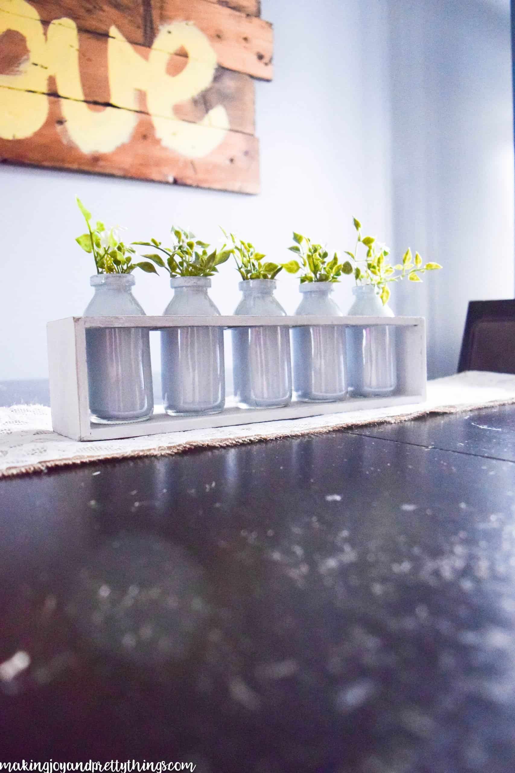 The completed summer centerpiece sits on a dark wood table, atop a lace and burlap table runner. A white chalk-painted box holds five white-painted glass jars, each holding sprigs of faux greenery and flowers.