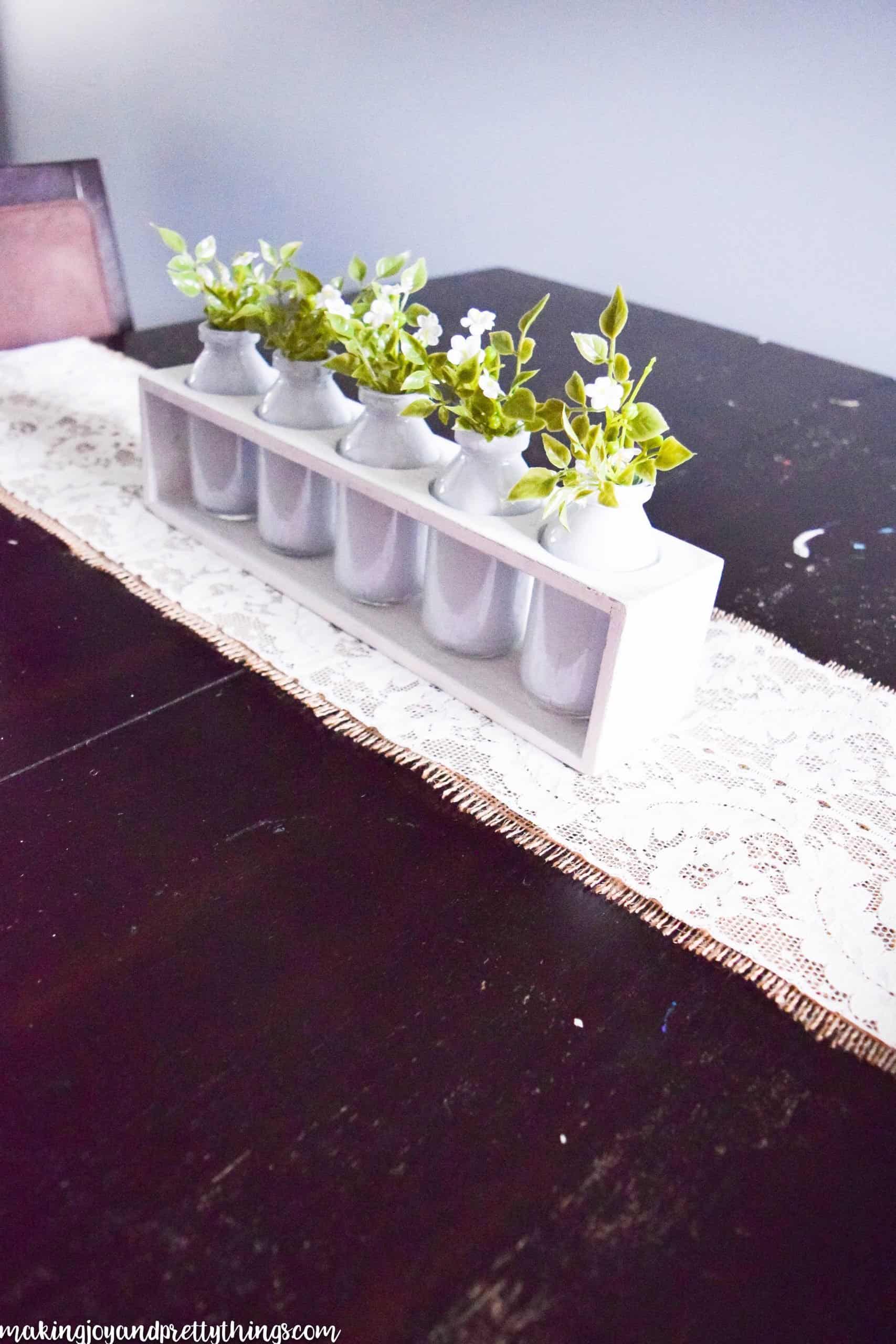 A side-angle view of the completed summer centerpiece and lace table runner. The centerpiece is made from a white wooden box with five holes in the top, each holding a white-painted glass jar filled with faux greenery and white flowers. The centerpiece sits on a lace and burlap fabric tablerunner.