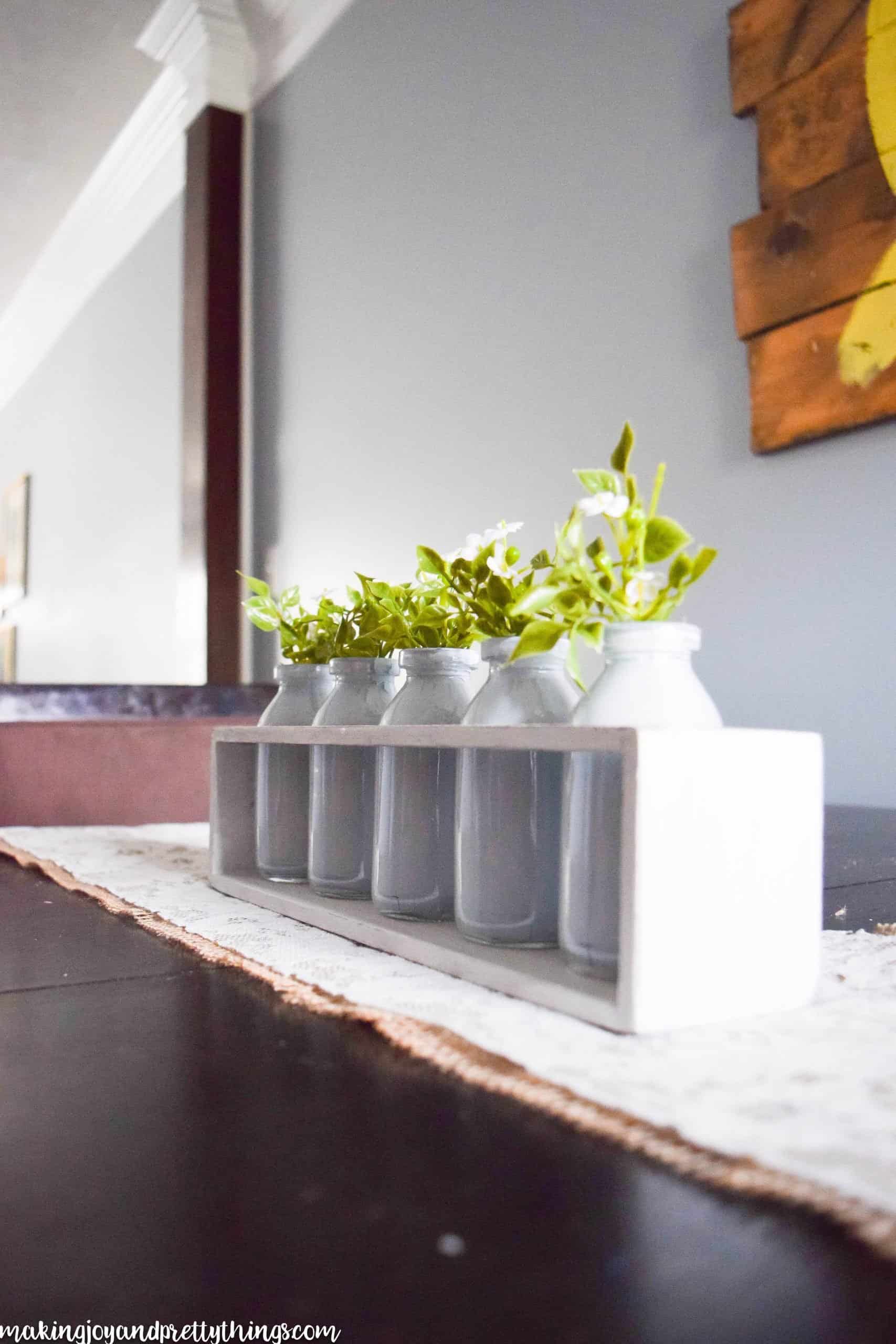 A side view of the summer centerpiece made from a white box, glass jars, and faux greenery. The centerpiece sits on a burlap and lace table runner in the center of a dark wood dining room table.