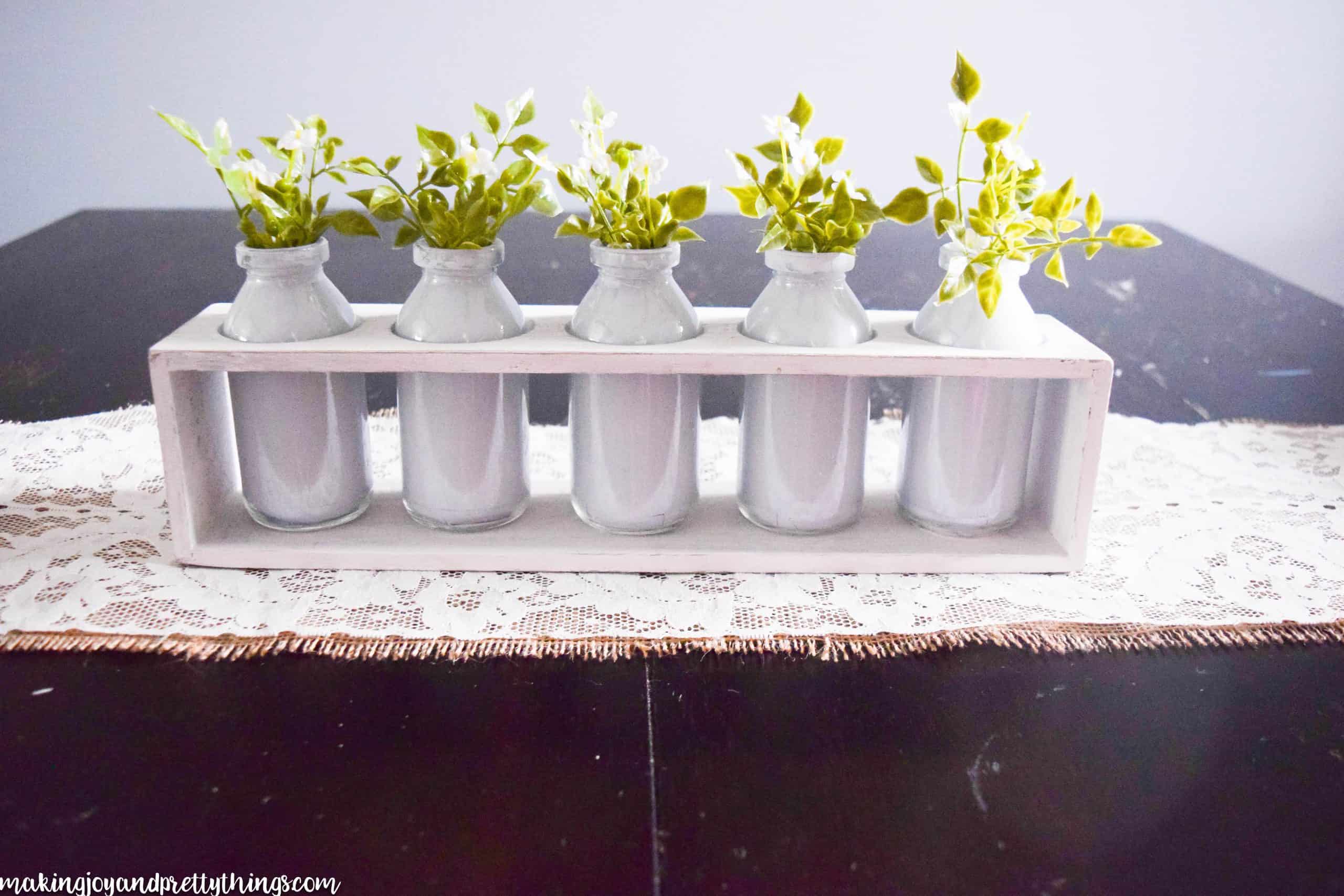 A full look at the completed summer centerpiece, sitting on a dark wood table. A white-painted wood box holds five white-painted glass jars, each filled with faux greenery. The centerpiece sits on a homemade lace and burlap table runner.