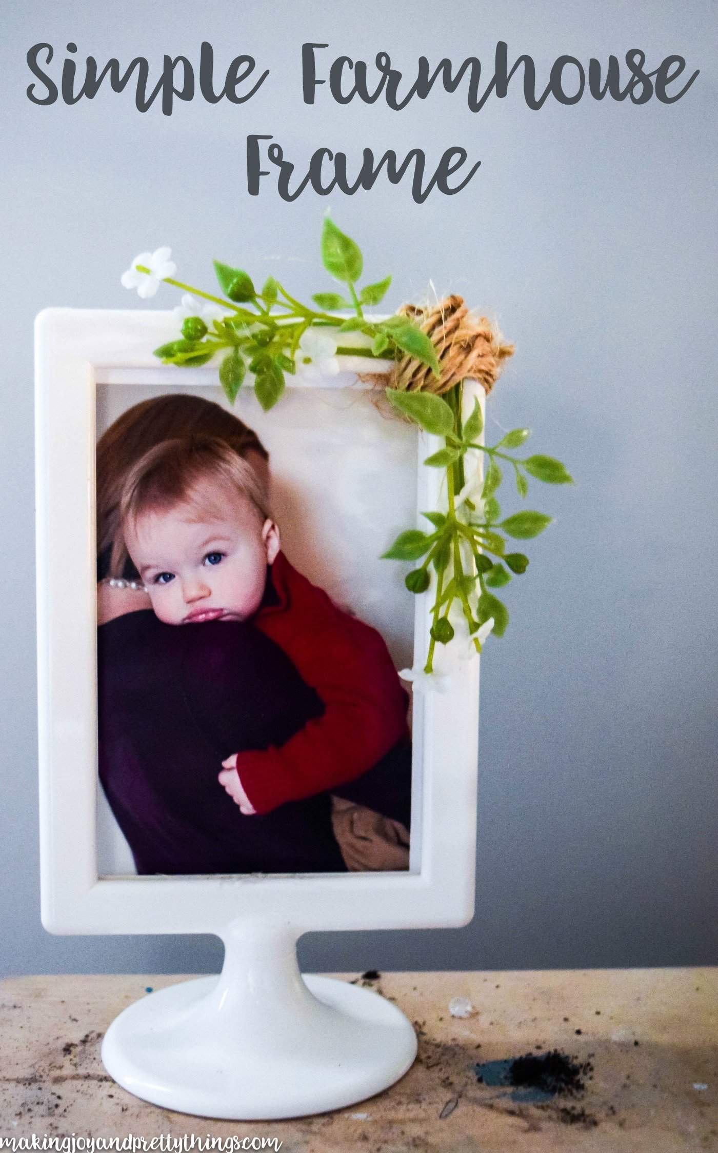 A white picture frame with a round base sits on a wood surface in front of a light blue wall. Inside the frame is a picture of a mother holding a toddler with big blue eyes, looking over the mother's shoulder. Sprigs of faux greenery and small white baby's breath flours and a string of twine are glued to the frame. Black text overlay on the image says "simple farmhouse frame"