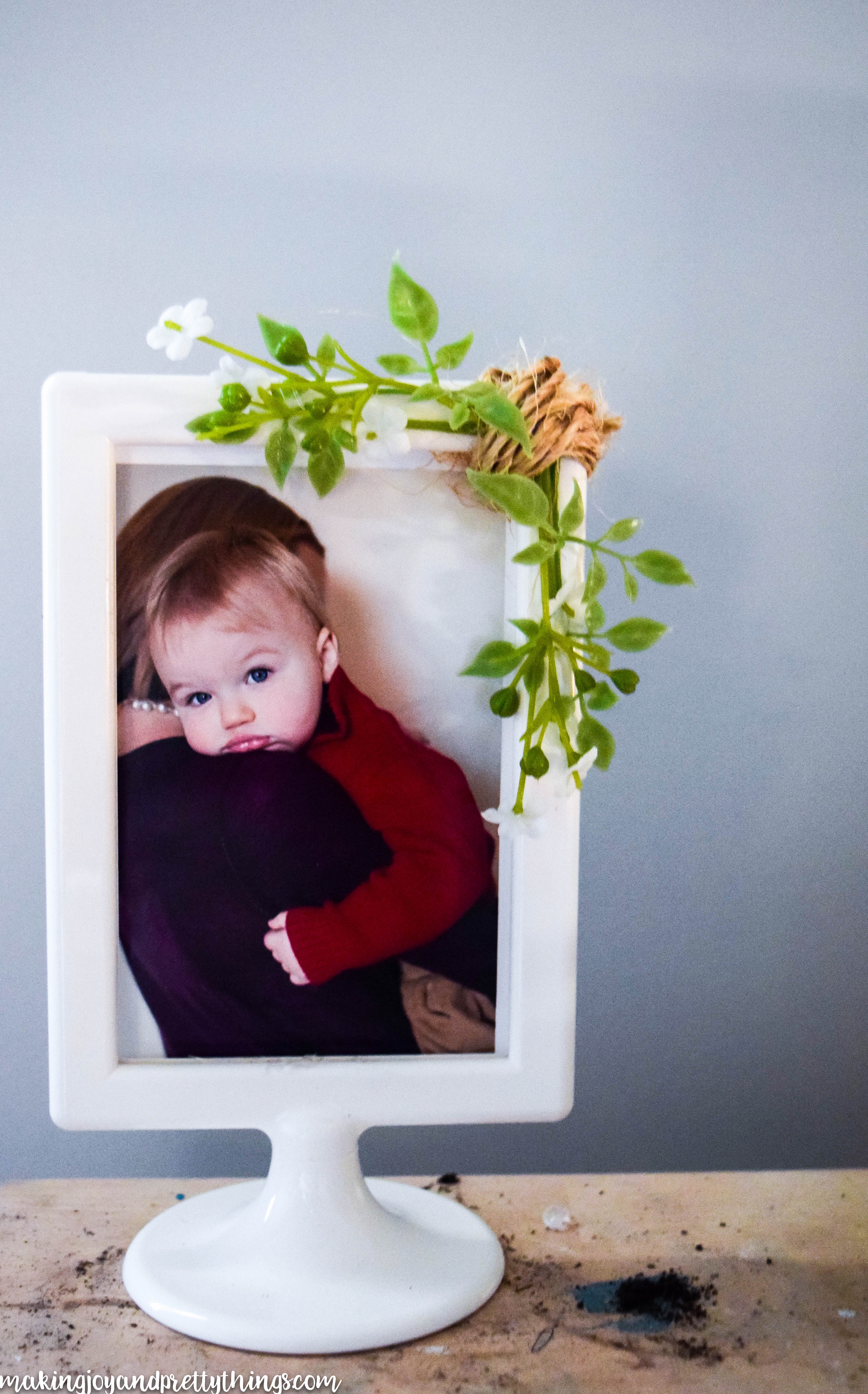 A simple white picture frame with a round base holds an image of a toddler looking over a mother's shoulder into the camera. Faux sprigs of greenery and small white flours are attached to the corner of the frame with crafting twine.