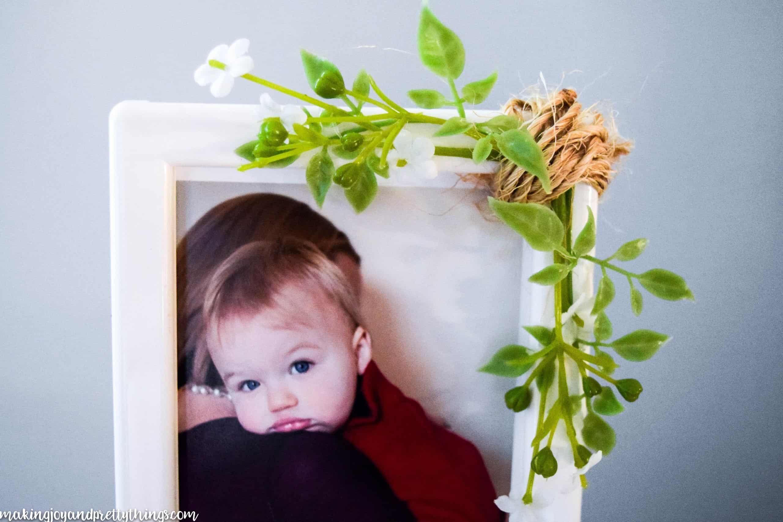 A simple white plastic picture frame is upgraded with a few sprigs of faux greenery and white flowers glued to the corner of the frame and wrapped with crafting twine. Inside the frame is a picture of a blue-eyed toddler resting their head on a mother's shoulder.