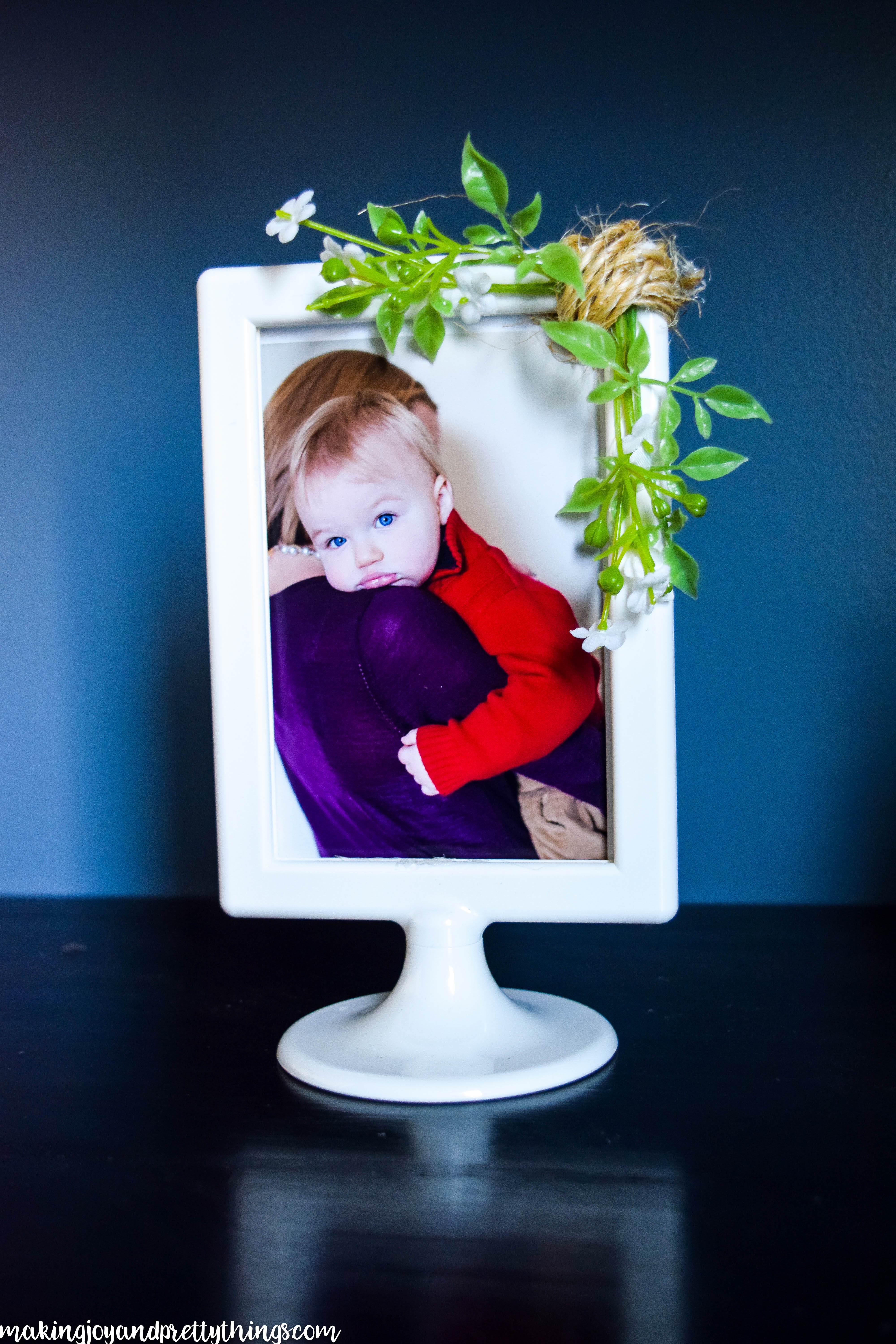 A simple white farmhouse frame with sprigs of faux greenery and small white flowers sits on a wood surface in front of a blue wall. The frame holds a colorful picture of a young toddler held in their mother's arms, looking over mother's shoulder directly into the camera.
