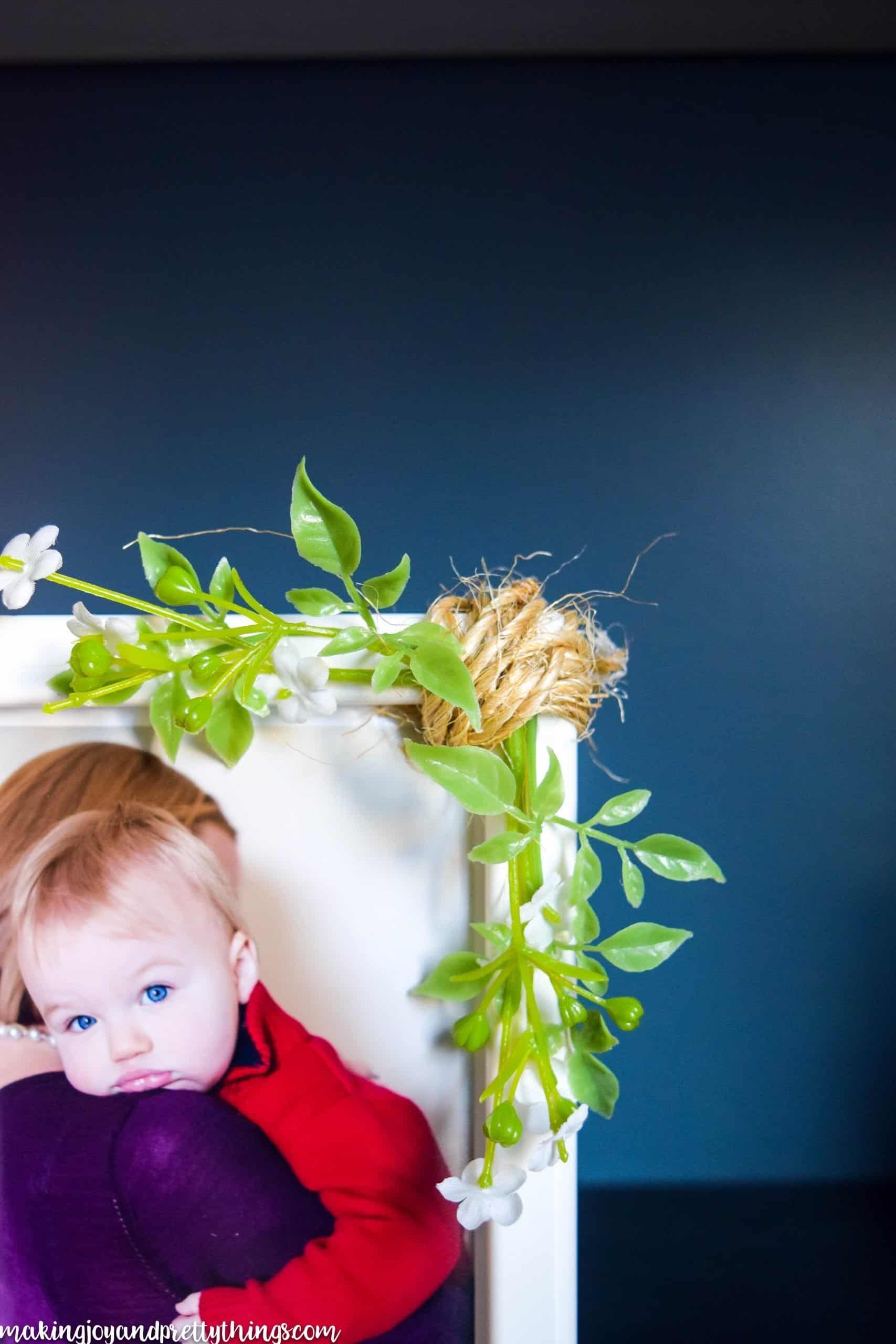 A close up look at the sprigs of faux greenery and twine attached the corner of a simple white farmhouse picture frame. Inside the frame is a picture of a toddler being held in a mother's arms.