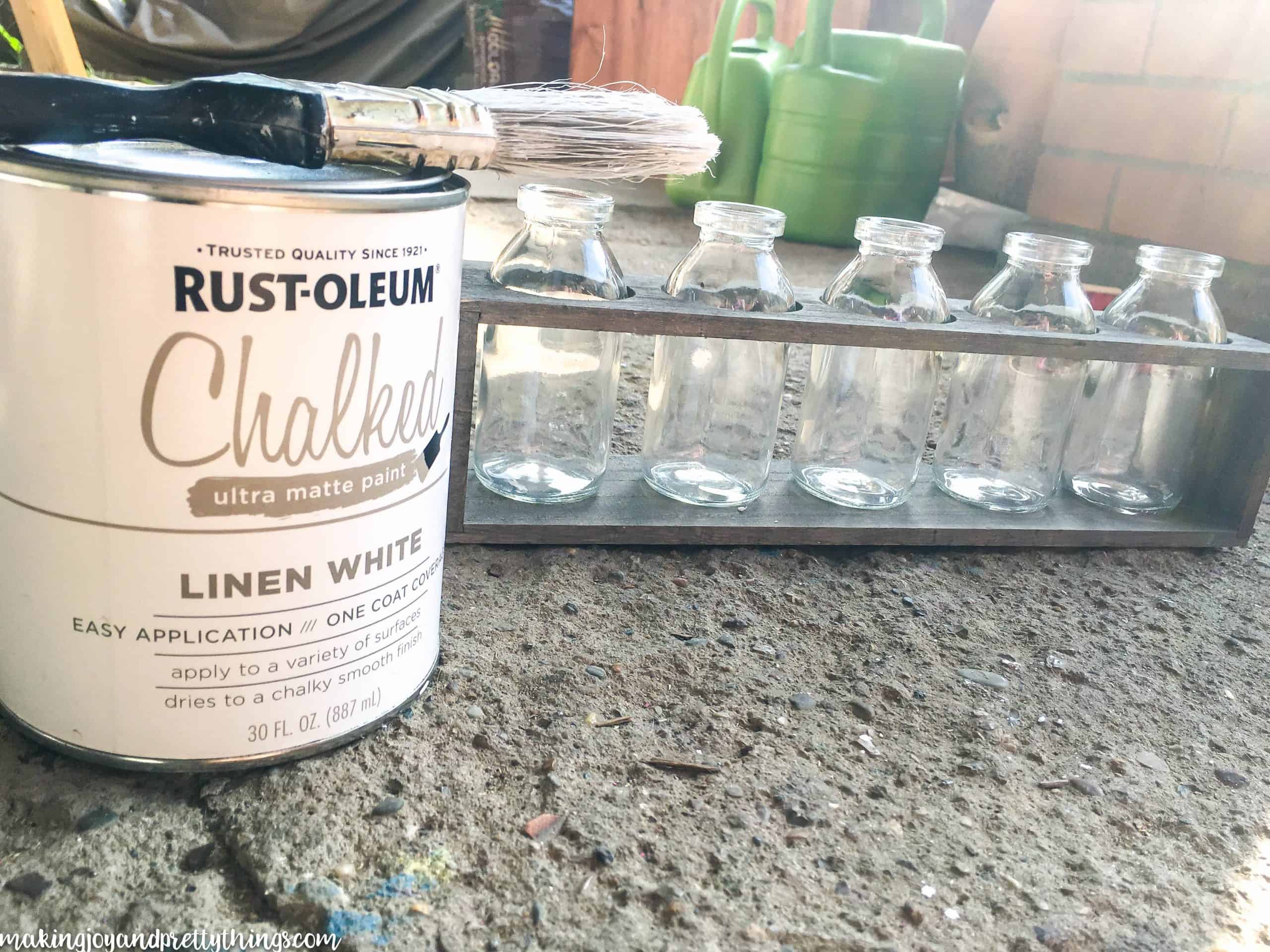 A can of Rust-Oleum chalked linen white paint and a wide paint brush sits next to a wood box with five glass milk jars.