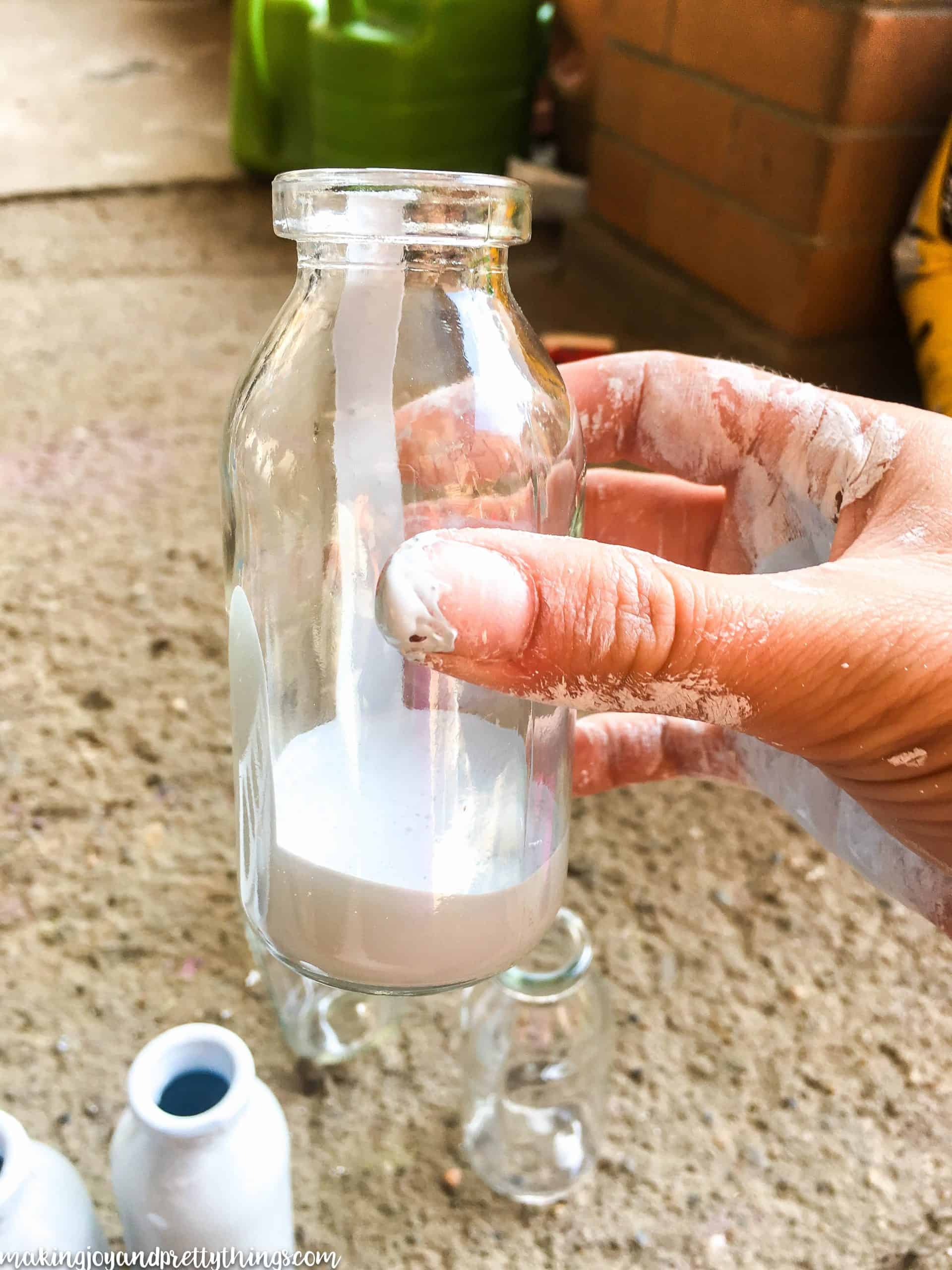 A small glass jar filled with an inch of white paint, and a woman's hand, covered in paint stains, holding the jar.