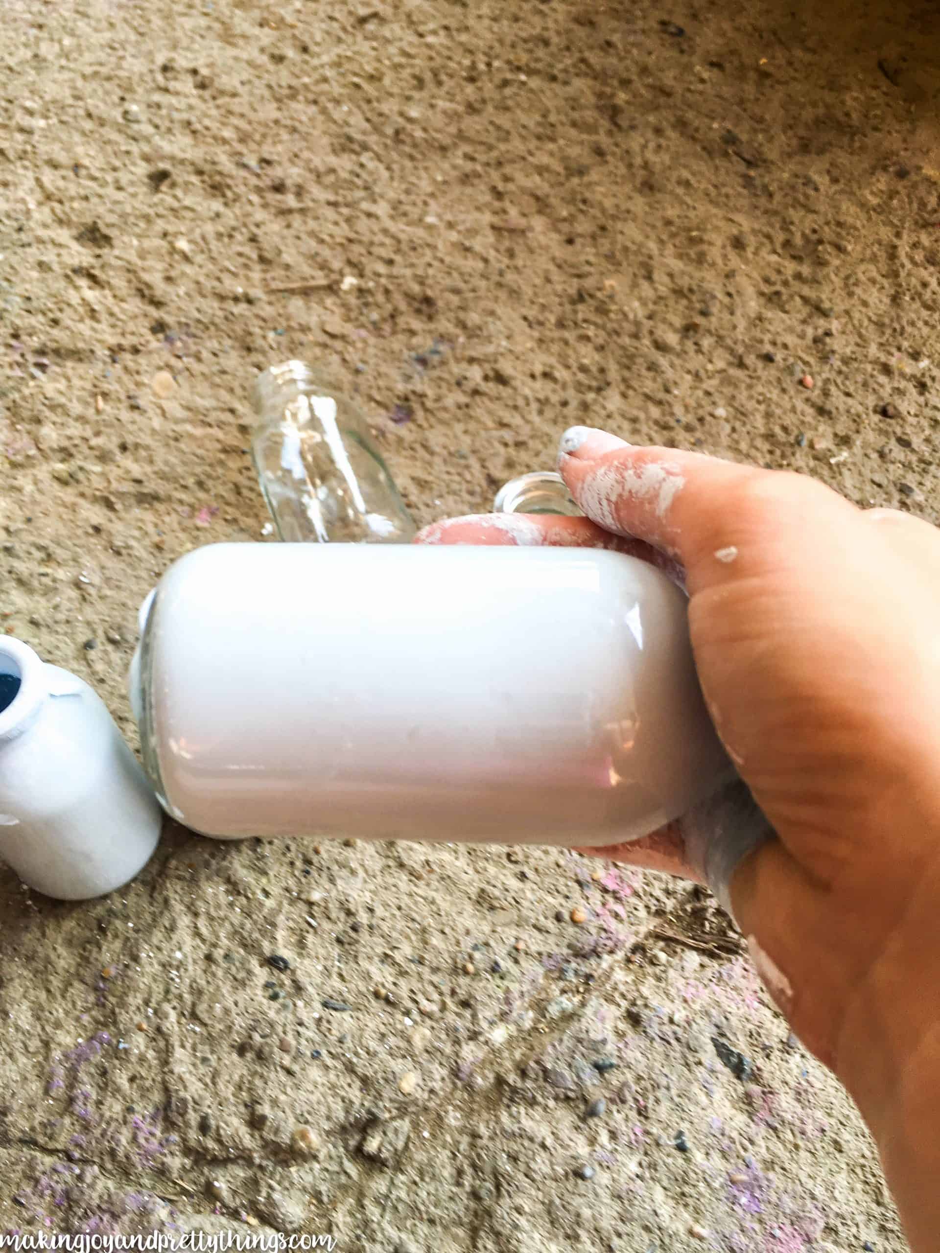 A paint-covered hand holds a glass jar, filled with white paint. The paint coats the inside of the jar, making it opaque.