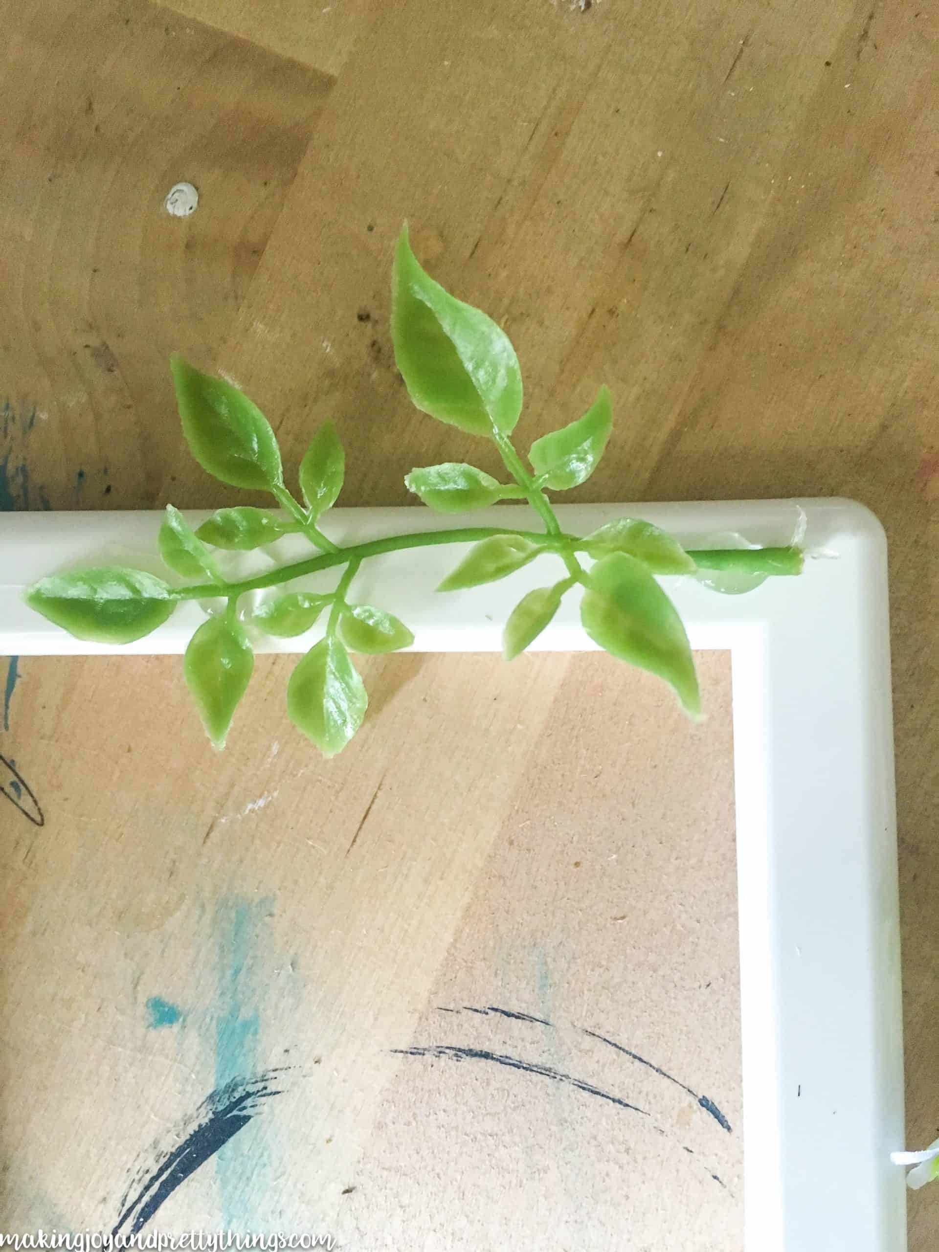 A single piece of faux greenery with small leaves is glued on to a white plastic picture frame.