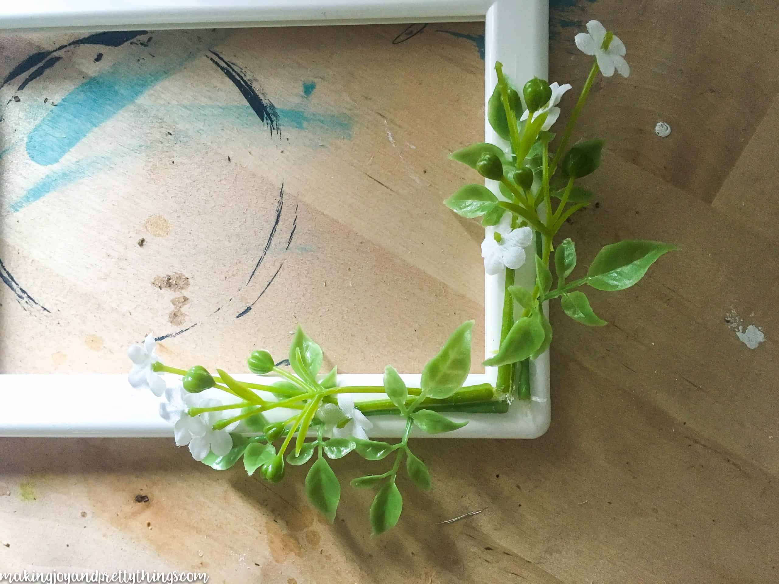 Two sprigs of faux greenery and small white flowers are glued on to either side of a corner of a white plastic picture frame.