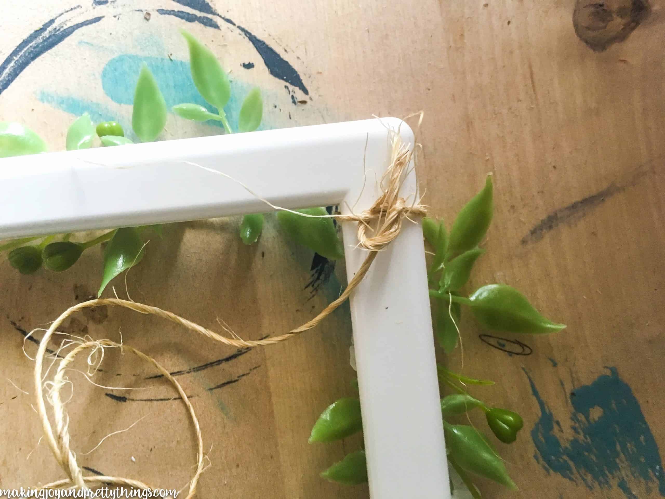 A long piece of craft twine is tied in a knot around the corner of a white picture frame. Faux greenery sprigs are visible, glued to the front side of the frame.